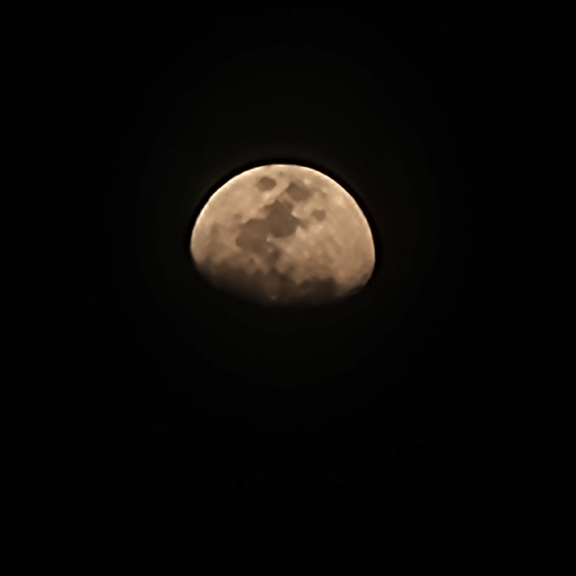 This is a close-up, slightly blurry photograph of the moon against a black, night sky. The moon is not fully illuminated; a portion of it is in shadow, suggesting a phase such as a waning gibbous or waxing gibbous, or possibly an eclipse. The visible surface of the moon displays craters and other topographical features, giving it a textured appearance. The lighting on the moon is warm, with hues of yellow and orange, hinting at atmospheric conditions or the color temperature of the light source. The overall impression is one of celestial observation, with the moon being the sole subject against the vast darkness of space. There are no visible people, objects, or text in the image. The time of day is clearly night.