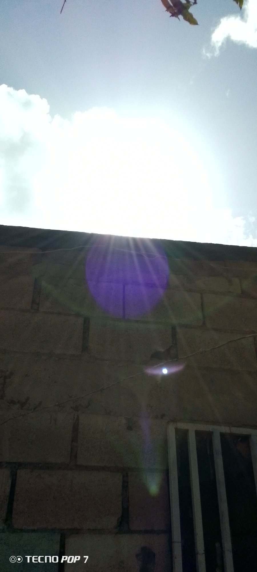 The image is an eye-level shot of a sunlit sky above a brick wall. The sun is bright and appears to be the primary light source, causing lens flare that manifests as a purple and white circle in the middle of the frame, with rays extending outwards. Some leaves, possibly from a tree, are visible at the top right of the image. The brick wall itself is roughly textured and appears to be part of a building. A thin wire runs horizontally across the wall. A white barred window structure is visible on the lower right side of the image. At the bottom left corner, a watermark reads "TECNO POP 7". There are no people or specific actions depicted. The location is identified as Altagracia, Venezuela.