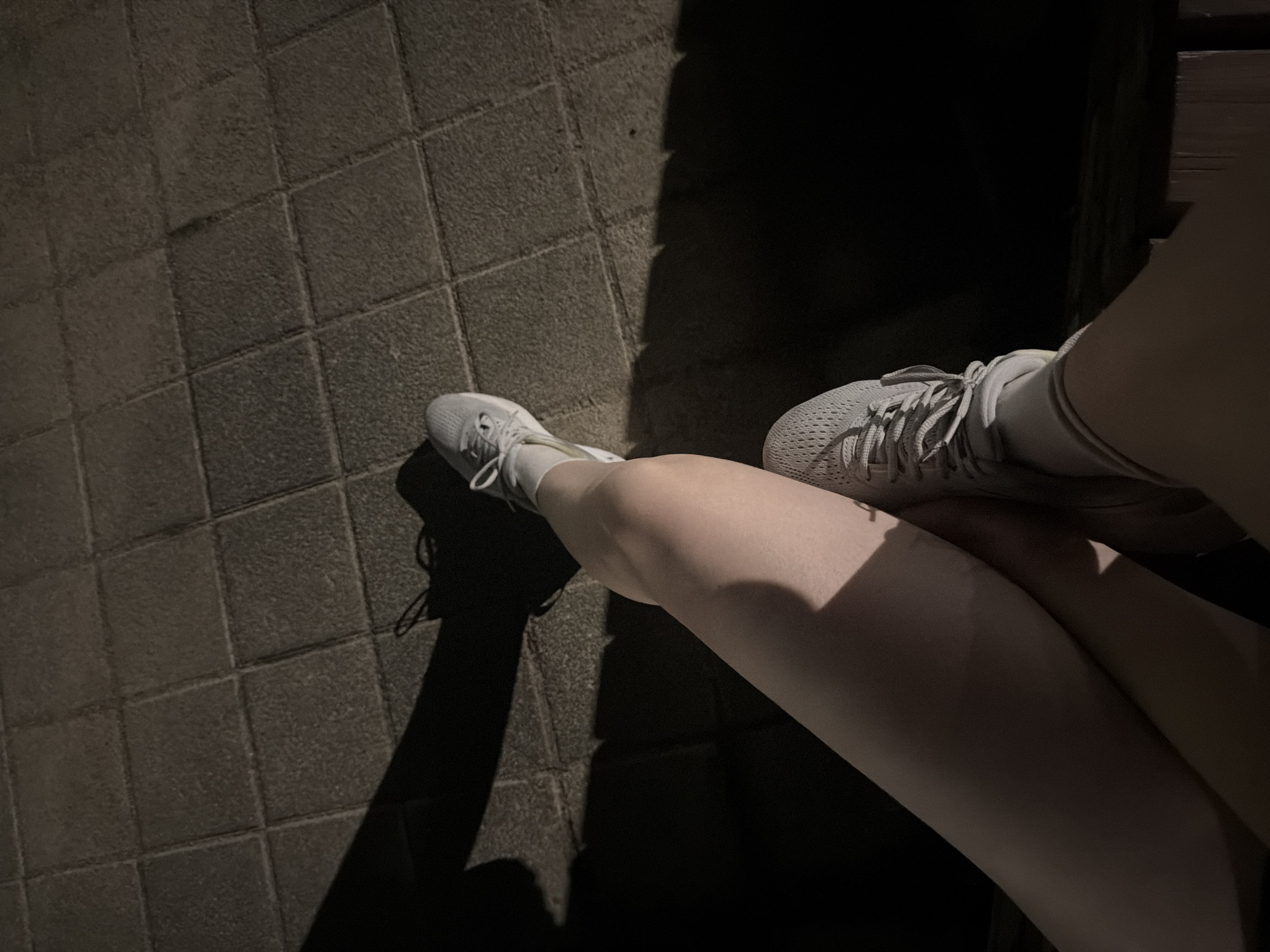 In Dongducheon-si, South Korea, a person is sitting on a wooden bench at night, casting a shadow on a tiled ground. The person's legs are crossed, and they are wearing gray athletic shoes with white laces. The scene is dimly lit, with the light source creating strong shadows.