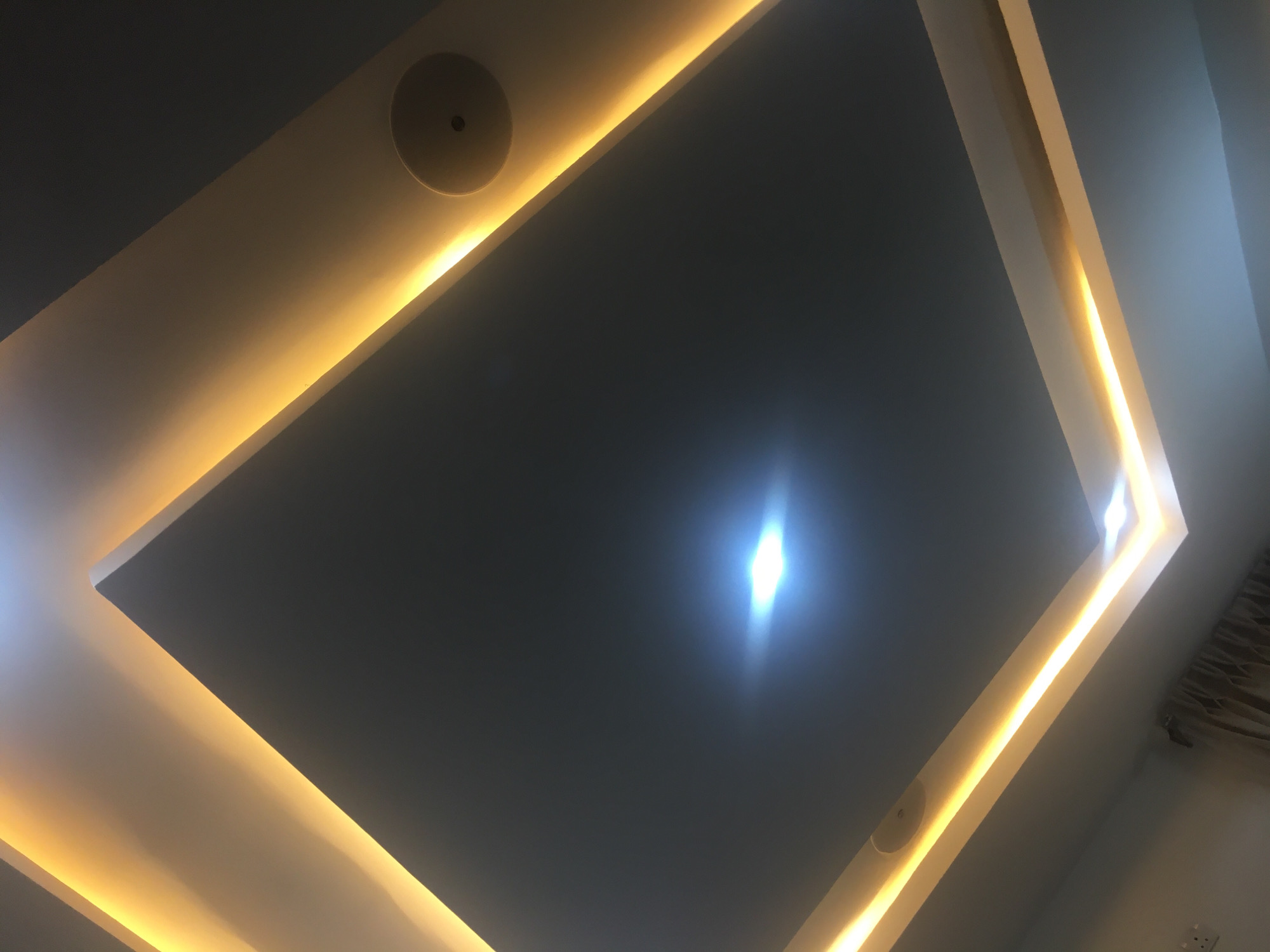 The image depicts a section of a ceiling with a recessed, square-shaped architectural feature. The ceiling is painted a dark gray color. A band of warm-toned lighting is installed within the recessed area, creating a glowing perimeter. A circular speaker is visible, embedded within the ceiling. The perspective of the shot is from below, looking upwards at an angle. There is a bright reflection in the center of the image from a lighting fixture. There are no people visible in the image. The location is unknown and cannot be identified from the provided visual information. There are no clues suggesting the location is Jos, Nigeria.