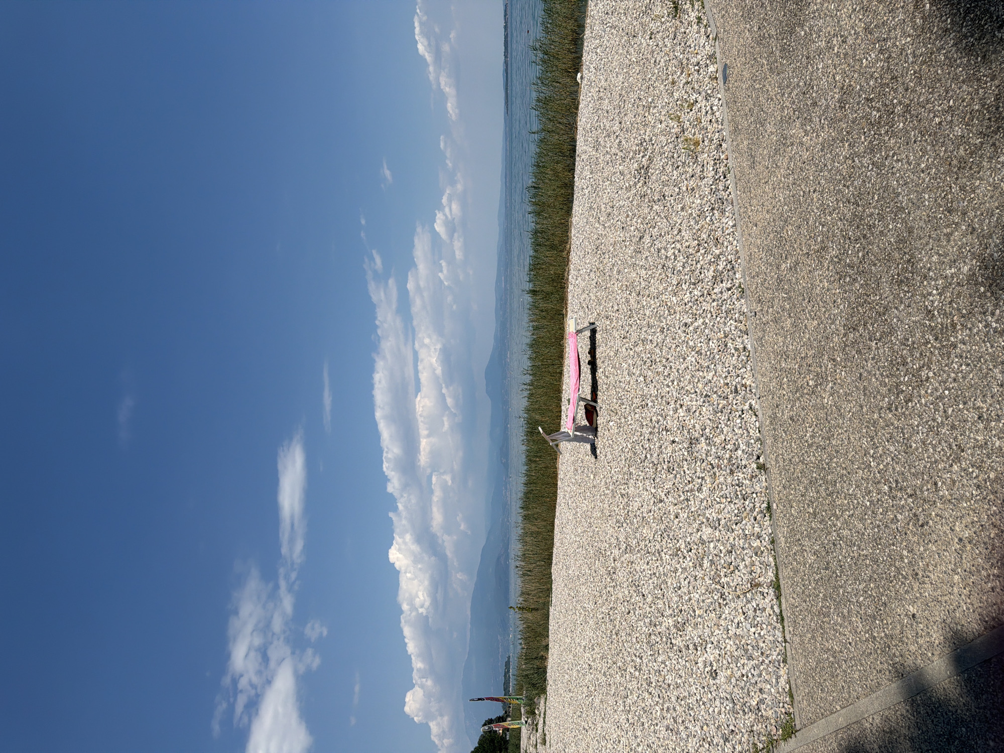 An image taken in Padenghe sul Garda, Italy shows a bright, sunny day at the lake. A pebbled beach area is in the foreground with a paved path to the left. A pink lounge chair is positioned on the pebbles near the waterline. Beyond the pebbles, tall reeds grow along the edge of a large body of water, likely Lake Garda. In the distance, hazy mountains form the horizon under a blue sky dotted with white clouds. A few colorful flags stand on the left side of the frame, near some trees. No people are visible in the scene.