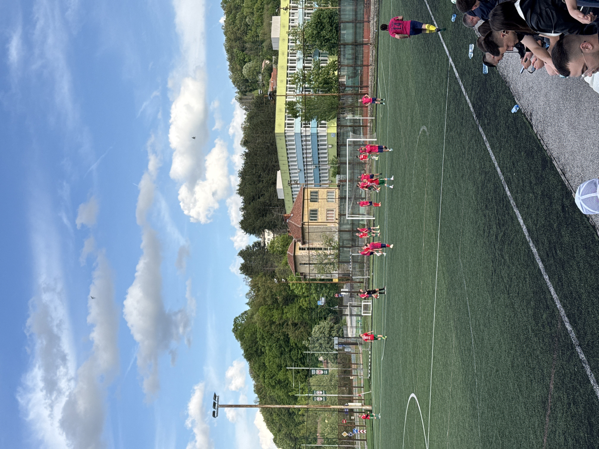 The image depicts a soccer field in Gabrovo, Bulgaria, with a team in red uniforms training. The players are spaced along the field, some appear to be jogging or stretching. Spectators are visible along one side of the field, near a paved area. Behind the field, there is a mix of buildings, trees, and a blue sky with scattered clouds. A bird can be seen flying in the sky. There are also street lights.