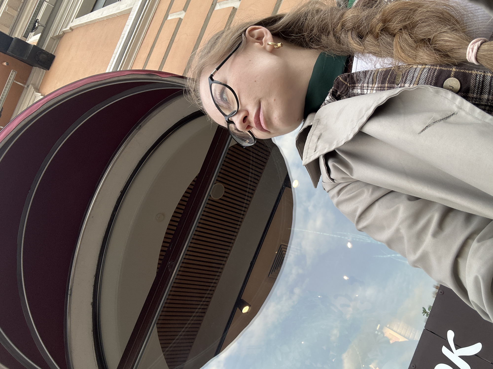 A woman wearing eyeglasses, a plaid shirt, and a trench coat stands near a window, with a burgundy and white striped awning above her. The window reflects a cloudy sky and the interior of a building with horizontal wooden slats and recessed lighting. Part of a sign with white lettering is visible in the lower right corner. The location is likely Saint Petersburg, Russia.