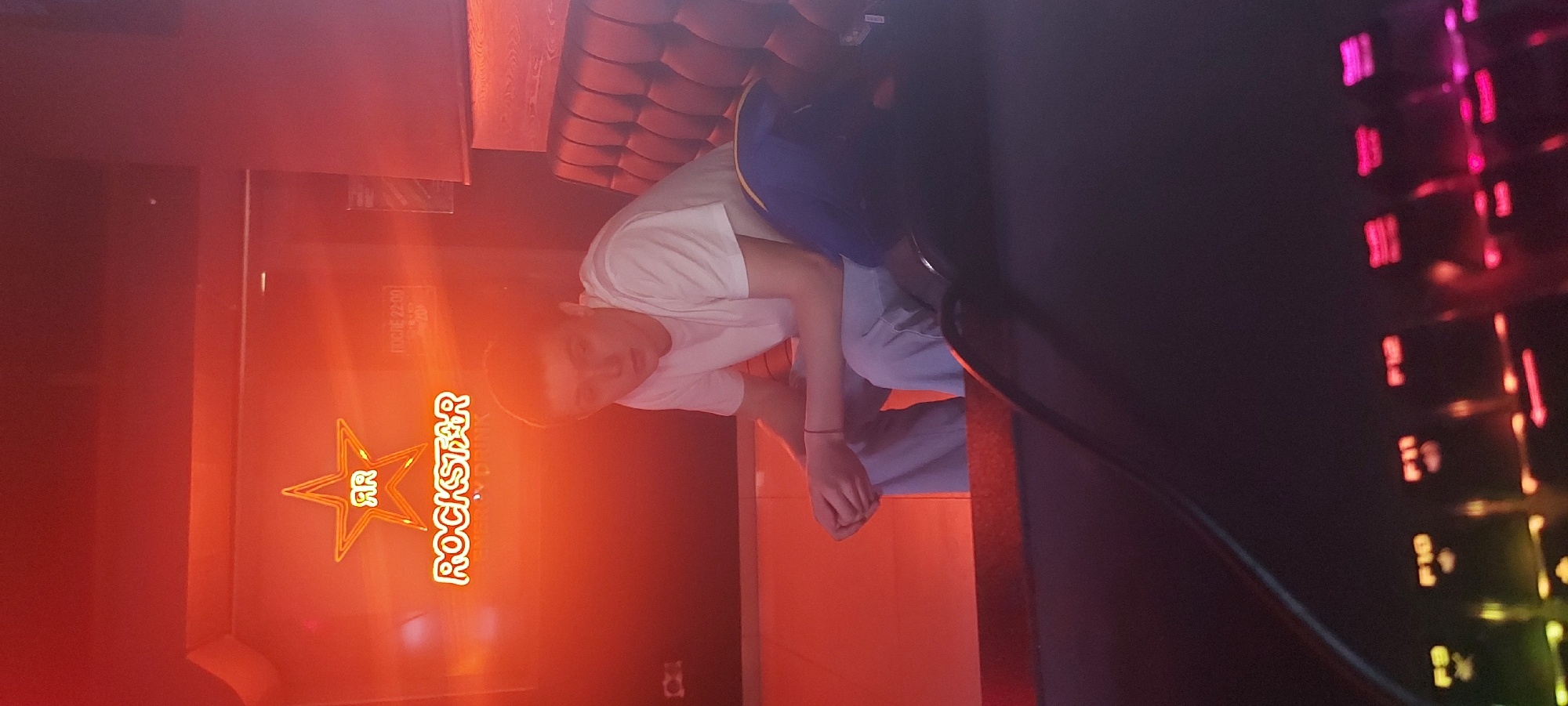 A vertical indoor shot presents a person sitting on a reddish-brown couch, illuminated by the glow of a "Rockstar Energy Drink" neon sign. The person wears a white t-shirt and light blue pants. To the right of the frame, a keyboard with multi-colored lights can be seen. Information on the Rockstar Energy Drink sign mentions Tashkent, Uzbekistan.