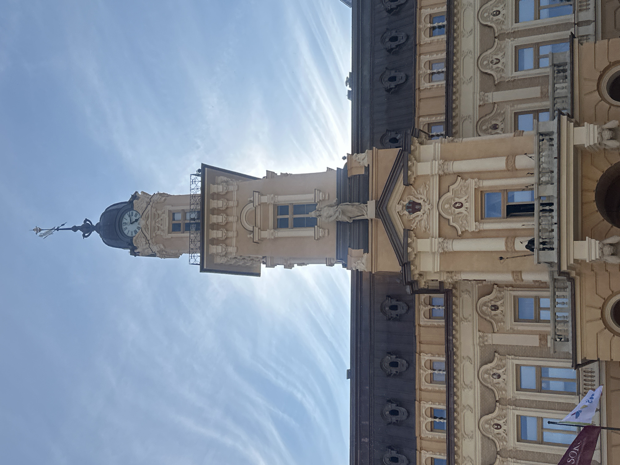 The image shows the facade of the historic Town Hall in Nowy Sącz, Poland. The building is constructed in a light-colored stone and features ornate architectural details, including columns, sculpted figures, and decorative window surrounds. A prominent clock tower rises from the center of the building, topped with a weather vane. The sky above is a bright blue with wispy white clouds. In the foreground, a balcony is visible with a single person standing on it, looking out. Flags are also present on the left side of the building, and some architectural elements suggest it is daytime. The overall impression is one of historical significance and architectural grandeur.