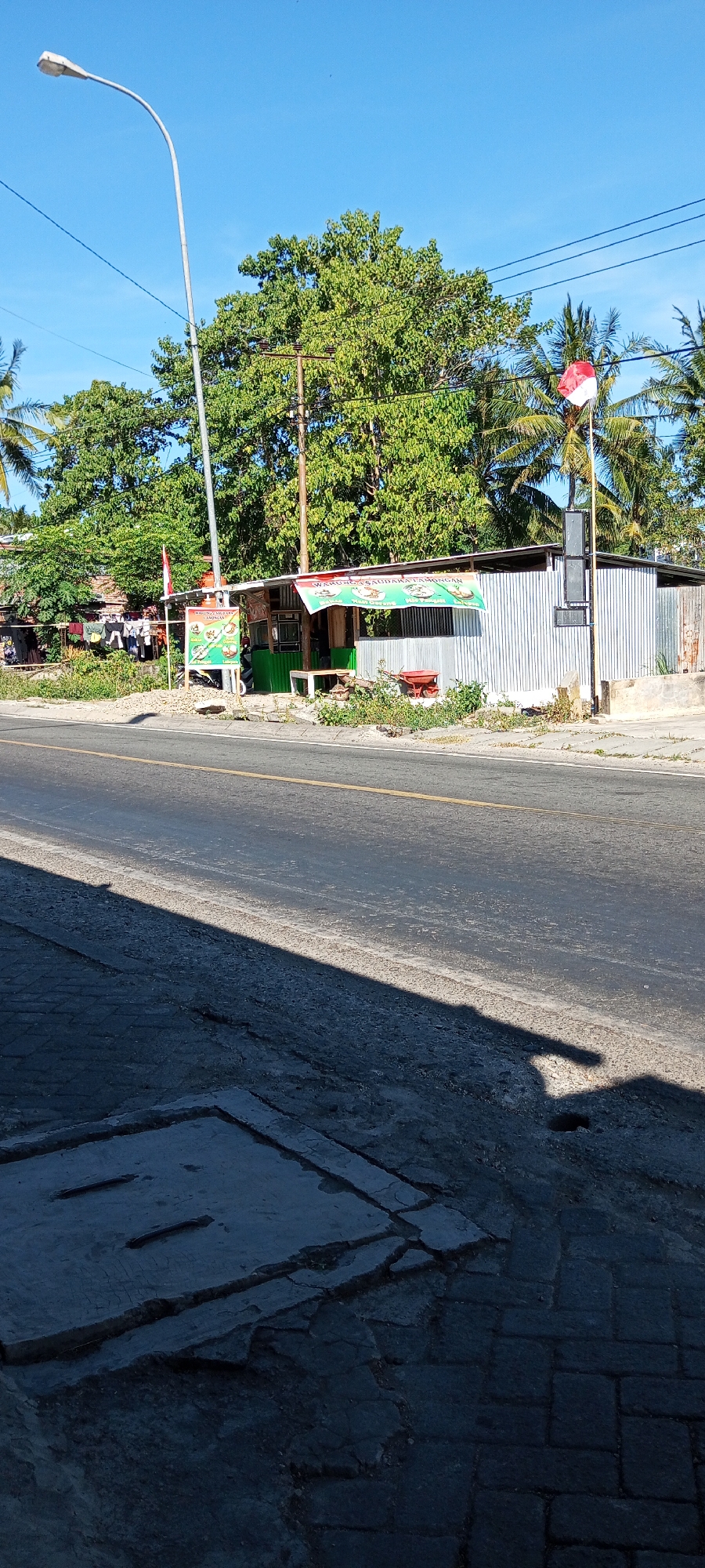 The image captures a scene in the city of Pakkanna, Indonesia. A roadside stall with a corrugated metal roof and siding is visible, adorned with a banner that reads "Warung Saudara Lamongan". Next to the stall, a flagpole flies the Indonesian flag. In the foreground, a paved road with a yellow center line stretches across the frame, partially obscured by the shadow of an unseen object. A concrete structure with two rectangular indentations is visible in the lower left corner of the image, partially cracked and surrounded by cobblestone pavement. Lush green trees and palm trees provide a backdrop to the scene. A street light stands tall on the left side of the image, and power lines crisscross the sky. The overall impression is a snapshot of everyday life in a tropical urban environment.