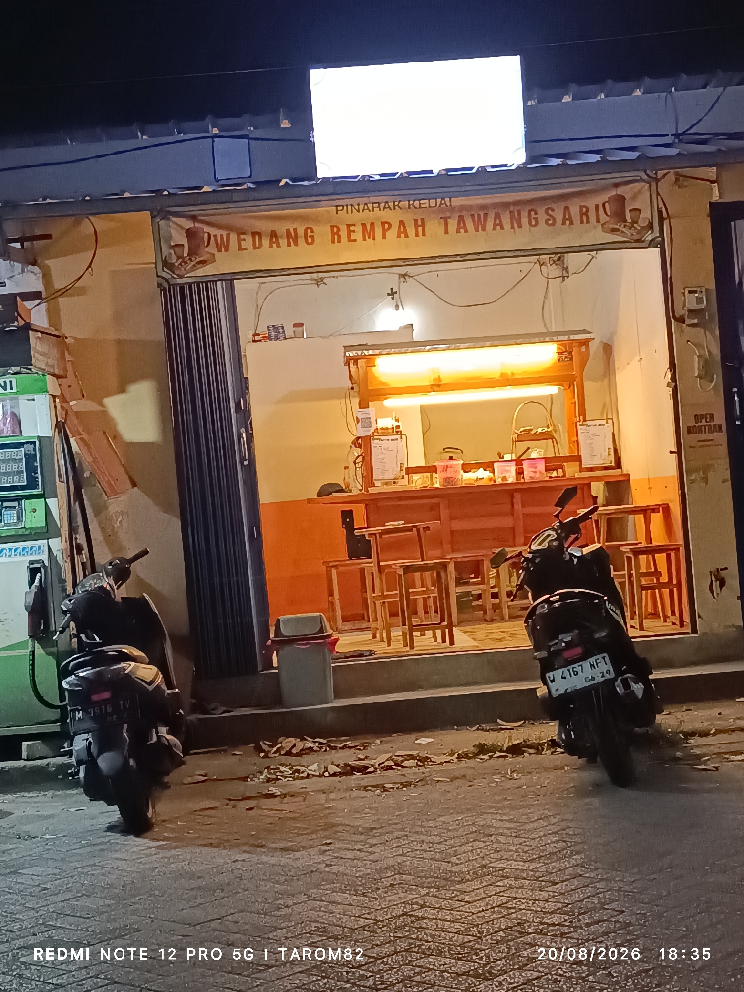The image shows the exterior of a small shop at night, likely a food or beverage stall, located on a paved street. The shop's name, "WEDANG REMPAH TAWANGSARI", is visible on a banner above the entrance. Inside, there is a counter with various items, illuminated by bright lights, and several wooden stools are set up for customers. A motorcycle is parked in front of the shop. To the left, part of a green fuel pump is visible, suggesting the proximity to a gas station. The environment appears to be an urban setting, as indicated by the presence of the fuel pump and the paved street. The date and time stamp on the image indicate it was taken on August 20, 2026, at 18:35. The presence of the motorcycle and the open storefront suggest the business is operational. The overall scene depicts a typical street-level business in Indonesia during the evening.