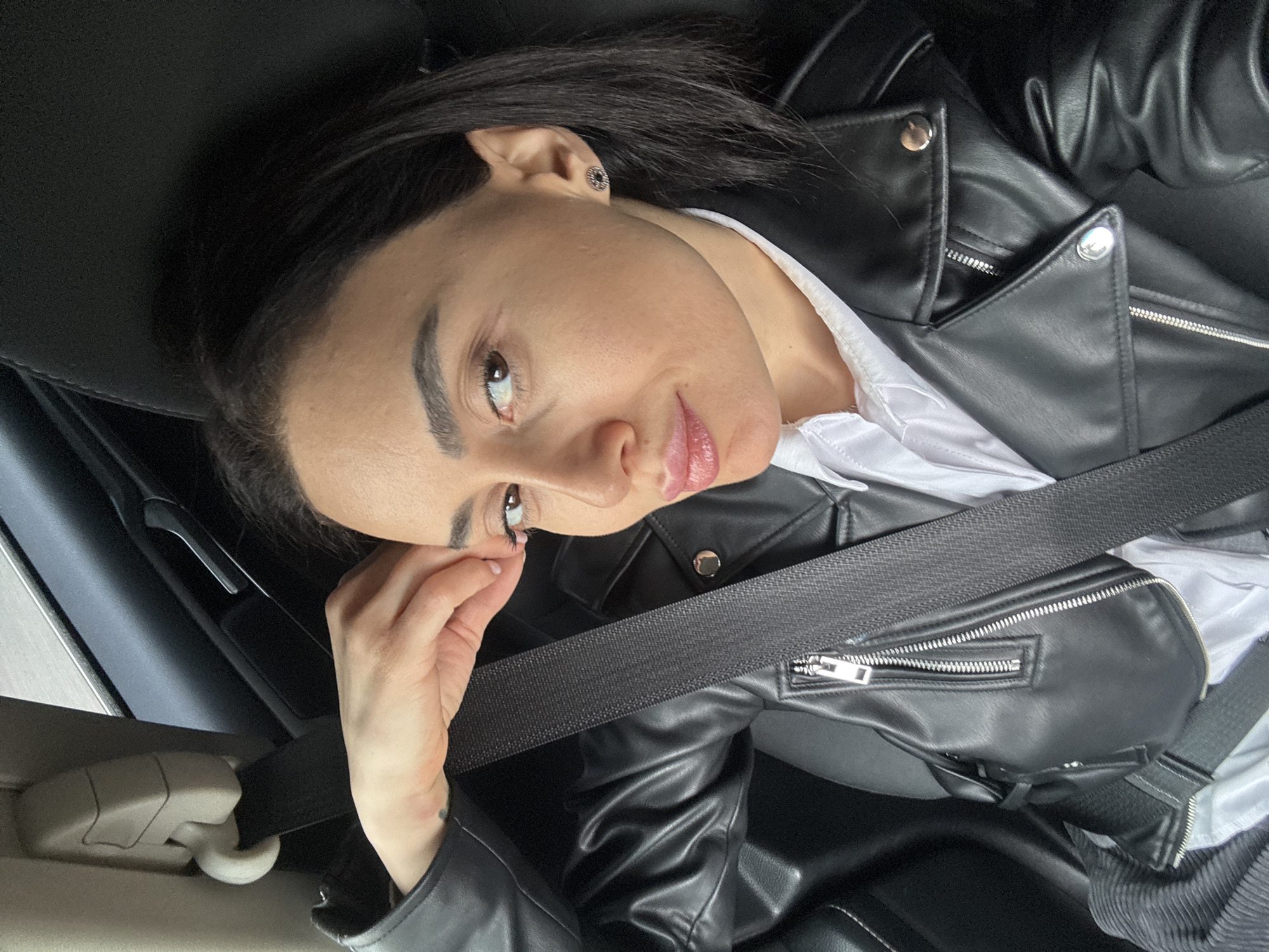 A woman with fair skin, dark hair, and light-colored eyes is seated in a car. She is wearing a black leather jacket over a white shirt, and a black seatbelt crosses her body. She has her hand near her eye. The interior of the car is black and light gray. The location is not discernible from the image.