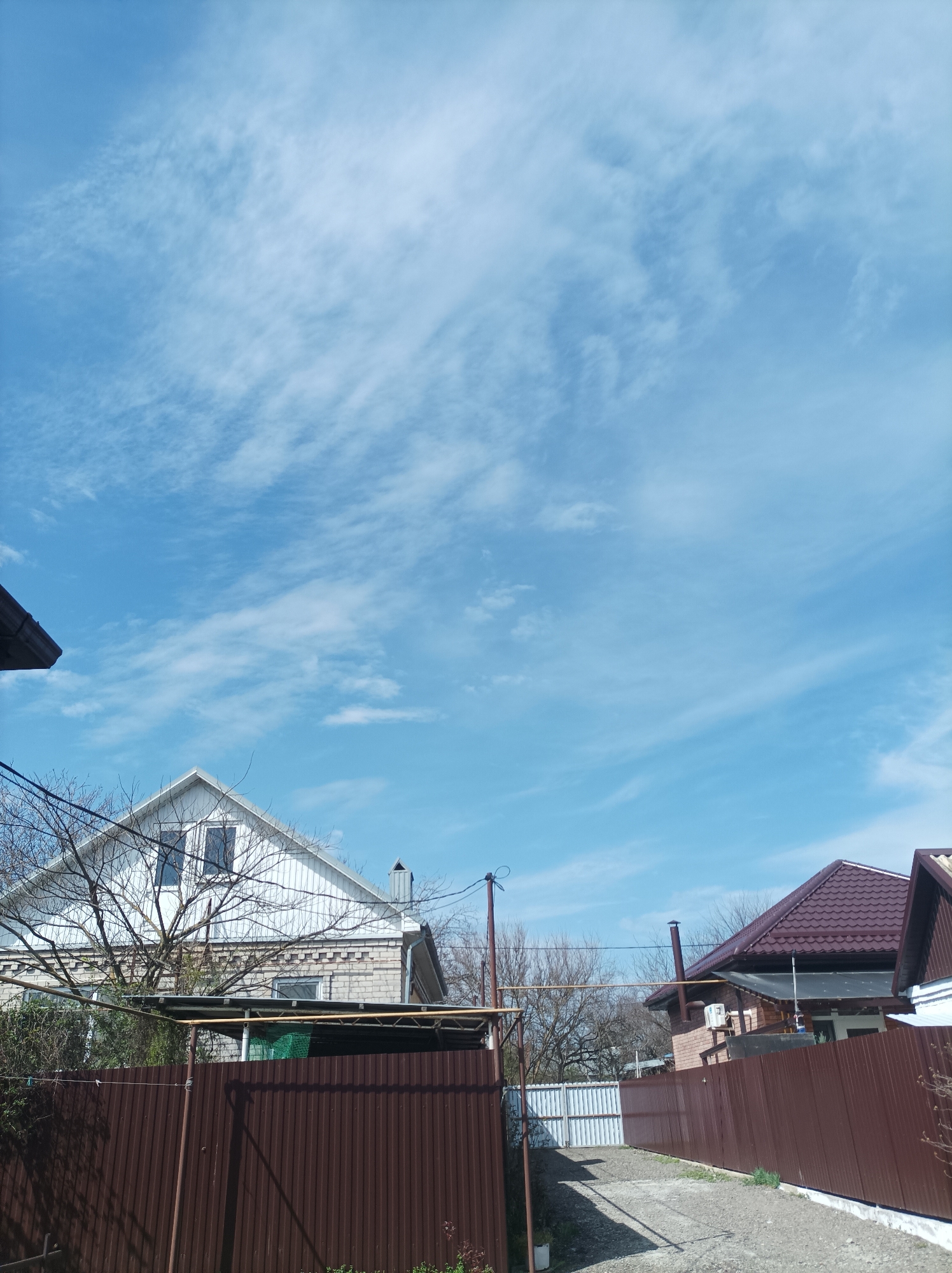 The image shows a neighborhood in Ilskiy, Russia under a bright blue sky with wispy clouds. Two houses are visible, separated by a narrow gravel driveway. The house on the left has a white gable roof with two windows on the top floor, a brick second story, and a brown fence in front. A leafless tree is partially obscuring the house. An awning or some kind of metal structure is attached to the front of the house. The house on the right has a brown tiled roof, a brick wall, and a similar brown fence along the driveway. Utility poles and wires are also visible overhead.