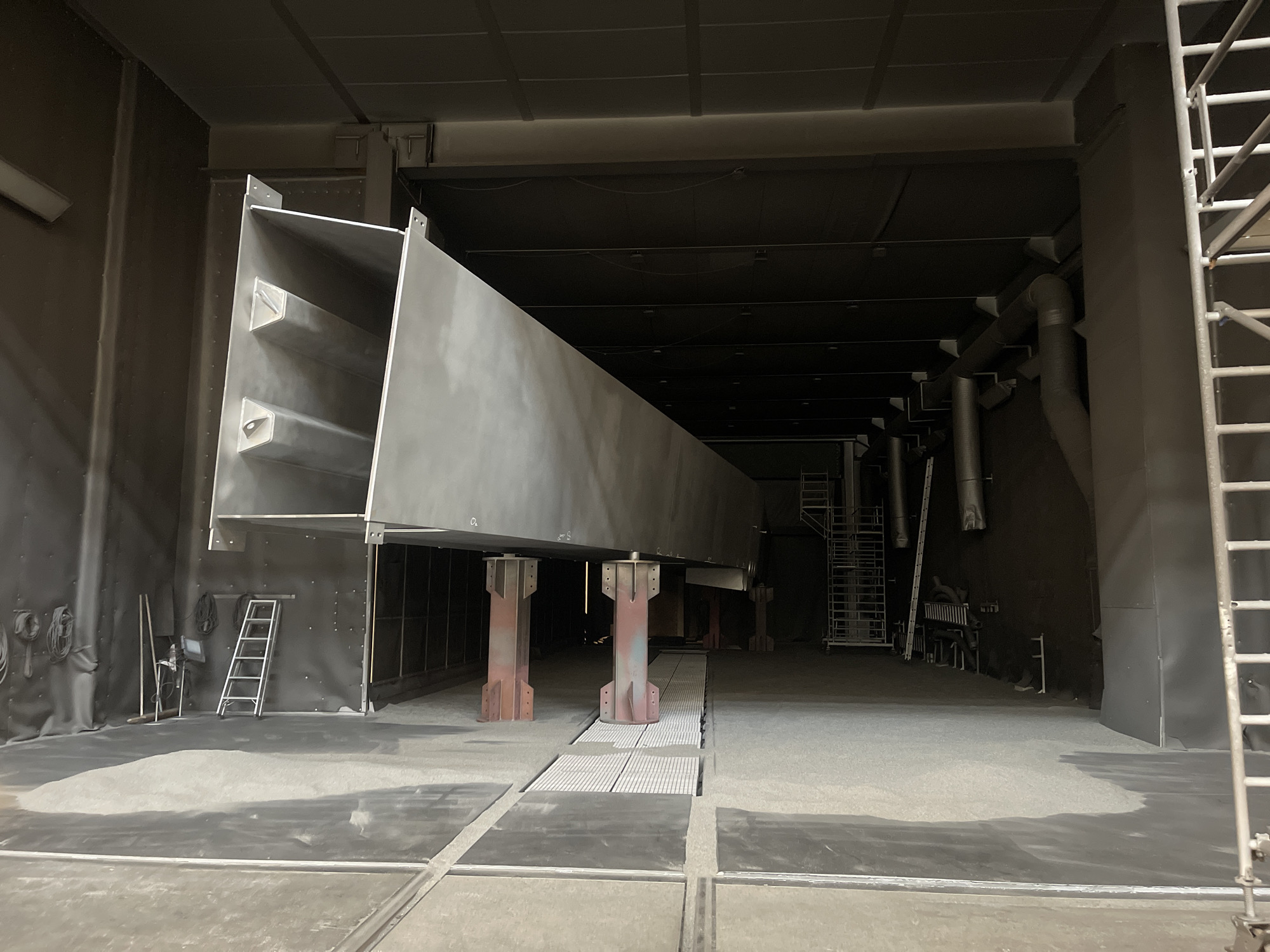 The image shows a large, silver-colored, metal beam suspended in a spacious, industrial setting. The beam is supported by three rust-colored pillars resting on a grey platform. In the background, there are metal staircases, ladders, and pipes. The walls and floor are a uniform shade of grey, suggesting a paint booth or similar industrial facility. Various tools and equipment are visible against the wall on the left side of the frame. The location is Unknown, Unknown.
