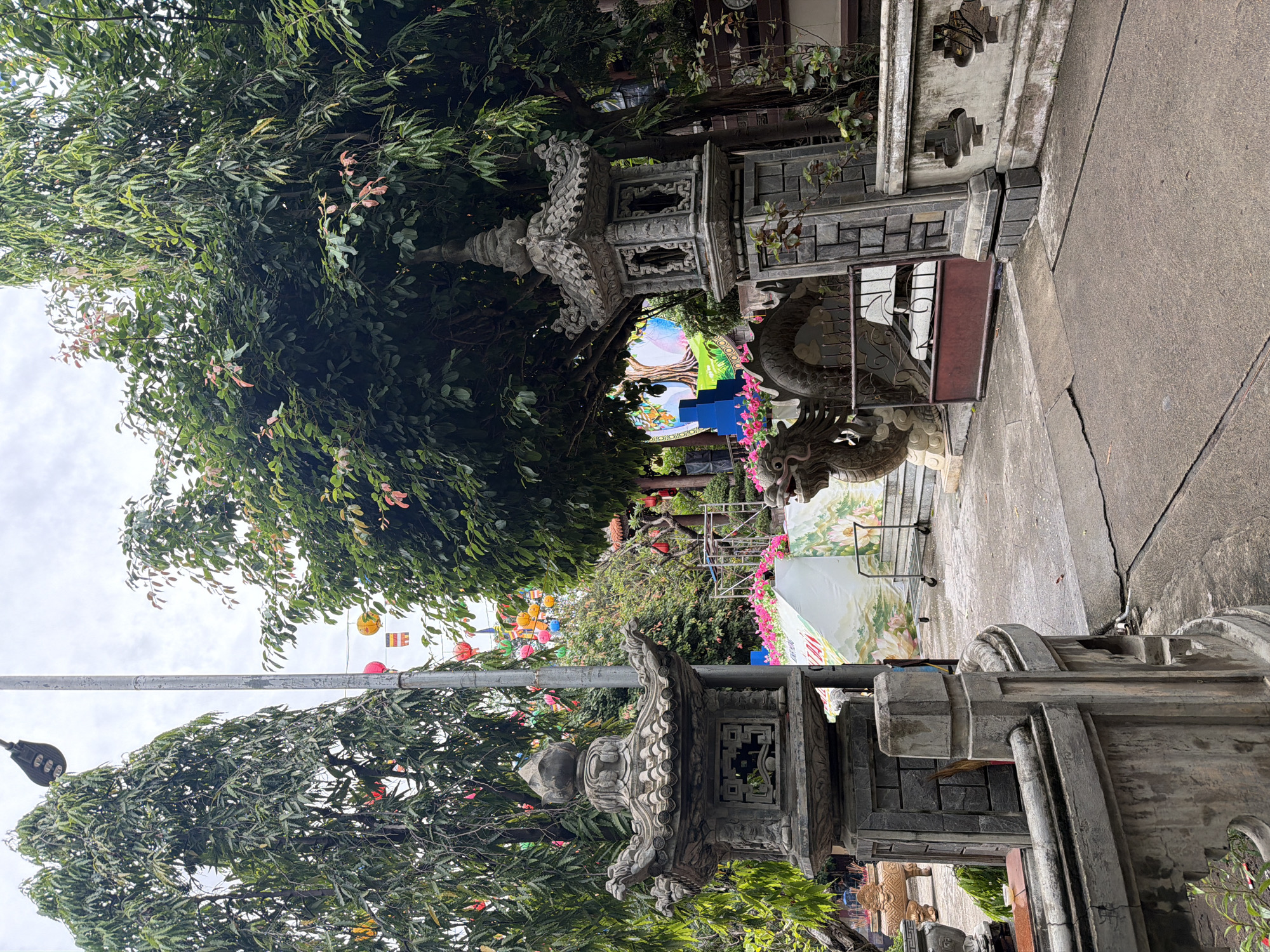 The image shows an outdoor scene in what appears to be a temple or religious site in Tay Nha Trang Ward, Vietnam. There is a stone lantern in the foreground. A walkway is present, leading towards an archway adorned with stone dragons. Colorful lanterns and banners are visible in the background. Lush greenery, including trees with green leaves, flanks the walkway. The sky is overcast. No people are visible in the frame.