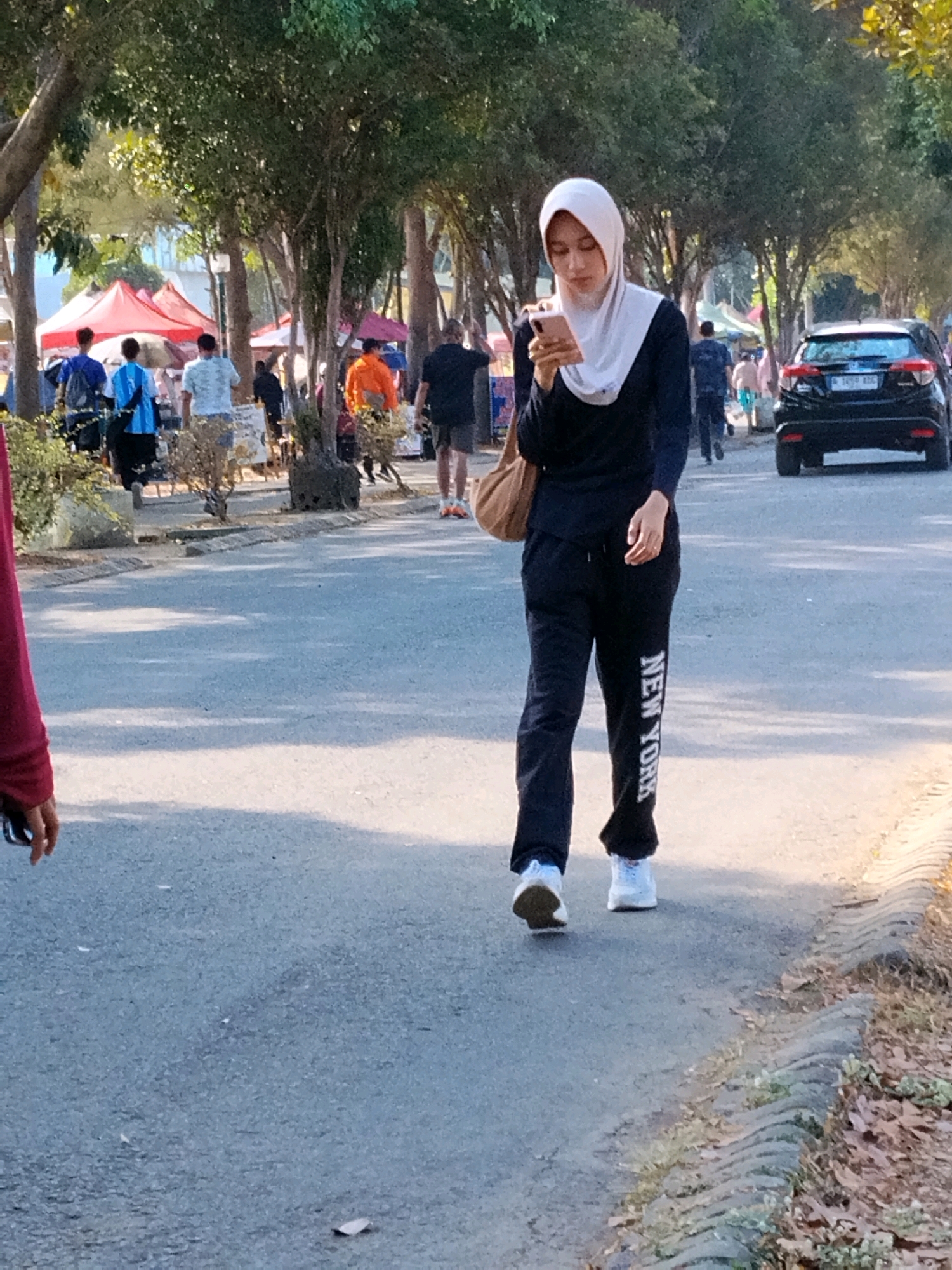 A woman wearing a light gray hijab and black activewear, including pants with "NEW YORK" written vertically on the left leg, walks on an asphalt path. She holds a smartphone in her right hand and a brown bag over her left shoulder. The path is lined with trees on the right and a grassy area with a curb on the far right. In the background, there are market stalls with red awnings, several people walking, and a black SUV parked on the right side of the street. The scene appears to be a public outdoor area, possibly a park or a street market, with a bright, sunny atmosphere casting shadows on the ground. The location is specified as Malang, Indonesia.