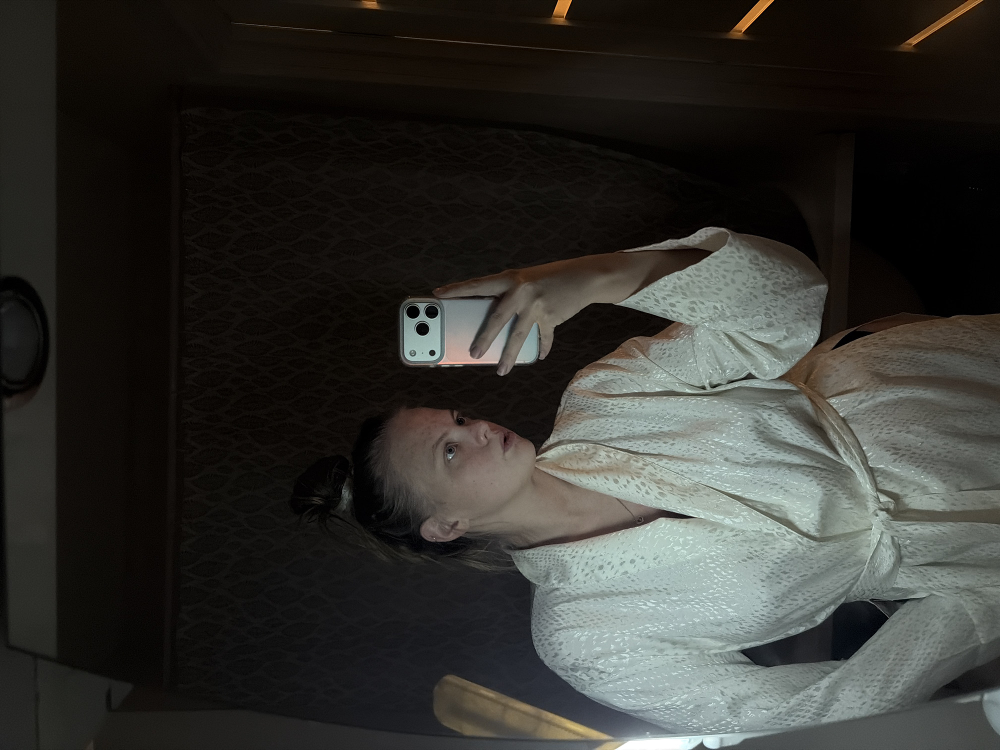The image shows a young woman indoors, in what appears to be a low-light setting, likely evening or night, in Izhevsk, Russia. She is wearing a light-colored, possibly cream or white, patterned robe with long sleeves, featuring a subtle, textured design. Her hair is pulled back, and she has a neutral, focused expression, looking upwards and slightly towards her smartphone.
She holds a smartphone, identifiable as an iPhone with a three-lens camera system, enclosed in a clear or translucent case, in her right hand. The phone's screen emits a soft, warm light that illuminates her hand and a portion of her face. This suggests she is actively using the phone, possibly taking a selfie, recording a video, or viewing content on the screen. A small necklace with a heart pendant is visible around her neck.
The background is dark, featuring a patterned, dark fabric or material, which might be a headboard or wall covering. Above this, horizontal wooden slats or trim are visible. On the far left, a circular object, possibly a light fixture, is partially seen against a lighter wall. A faint, glowing light source is also visible at the bottom of the frame. No discernible text is visible in the image.