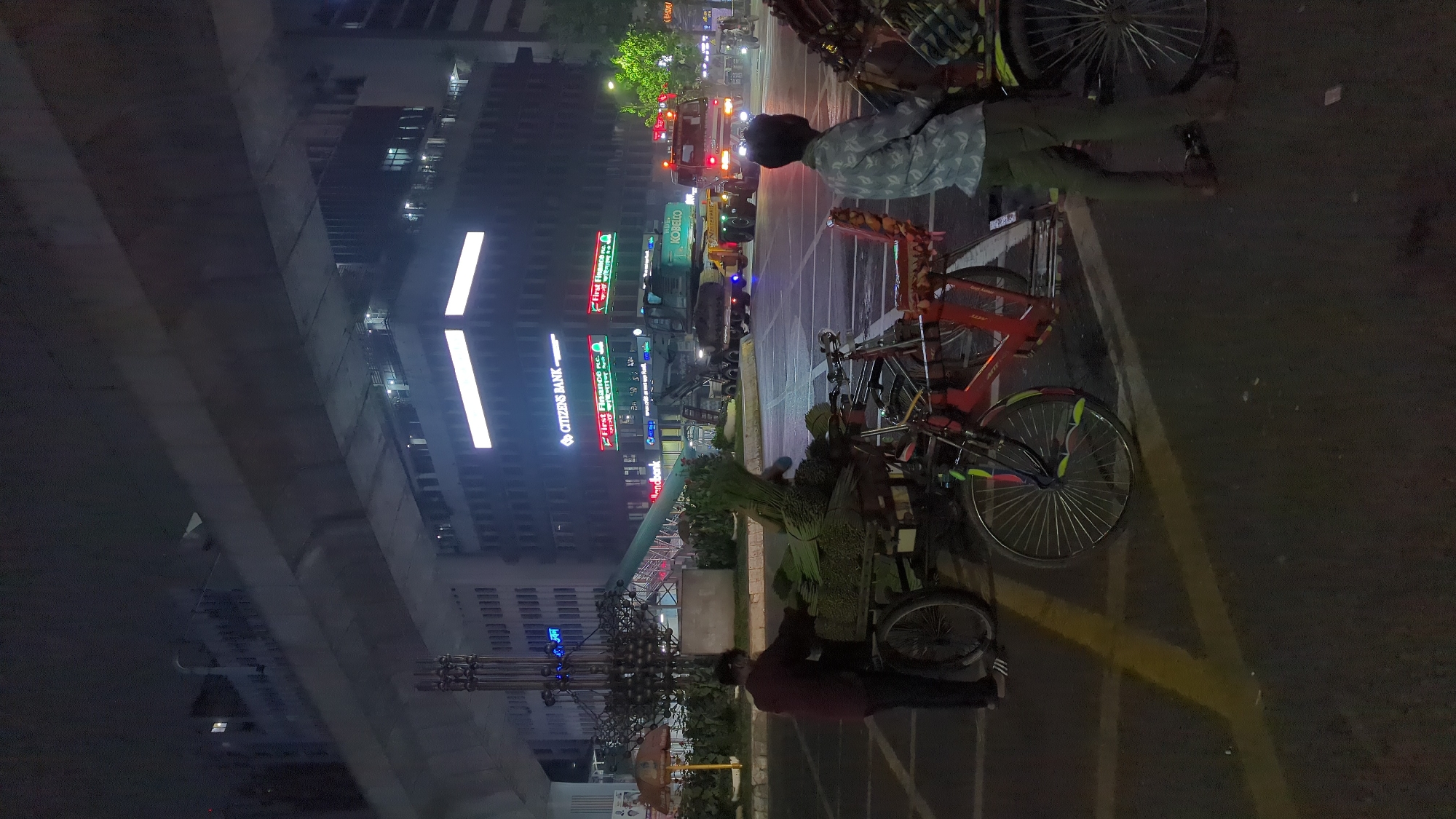 At night in Dhaka, Bangladesh, a street scene is captured with a mix of urban elements. A person is standing near a vendor's cart loaded with greenery, possibly flowers or leaves, while other individuals are nearby. Parked rickshaws line the roadside. The backdrop features a modern building illuminated with vertical light strips and neon signs for "Citizens Bank" and "First Finance", suggesting a commercial area. A large construction vehicle or crane is seen further down the street. Overhead, a portion of an overpass or elevated structure is visible. The setting appears to be a busy city street at night.