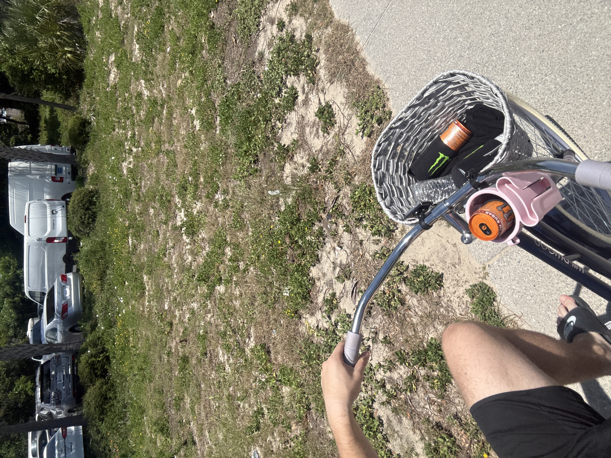 A POV shot from a bicycle handlebar in Panama City Beach, Florida, shows a person's hand gripping the handlebar and their legs and feet in black shorts and black slides. A wicker basket on the front of the bicycle contains a black t-shirt, a bottle of water, and an orange can of Monster Energy drink. A pink cup holder attached to the bicycle holds another orange can of Monster Energy drink. The bicycle is on a paved path, with a grassy area and several parked vehicles, including a silver sedan, a silver SUV, and white vans, in the background. The environment appears to be a sunny, outdoor location, likely a parking area near a beach.