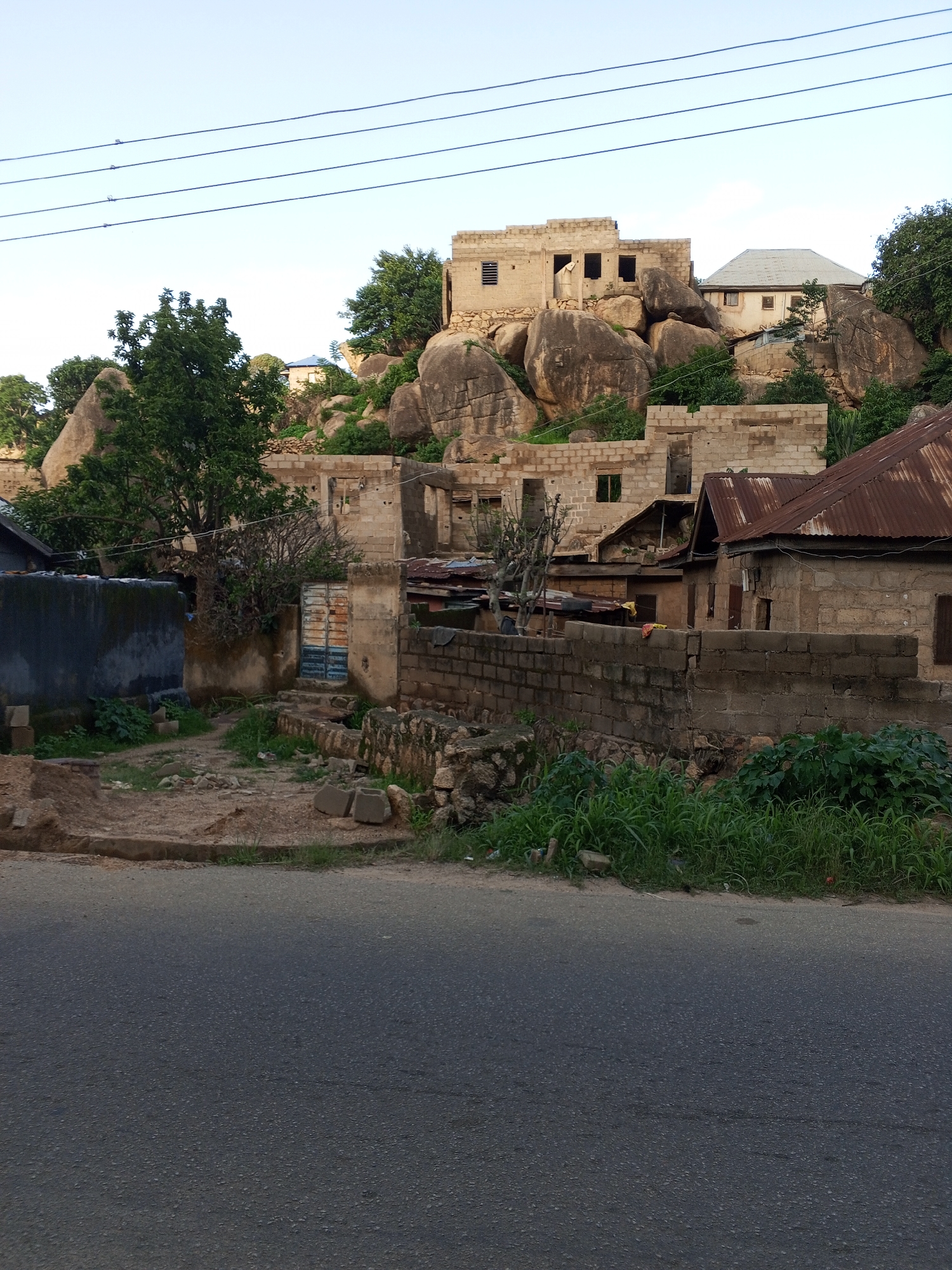 The image shows a view of a residential area in Kabong, Nigeria, with buildings constructed against a rocky hillside. In the foreground, a paved road runs horizontally across the bottom of the frame. Behind the road, there is a low brick wall. Beyond the wall, a yard area is visible with some scattered debris, including concrete blocks. Several unfinished or partially constructed buildings made of concrete blocks are present.
Further into the scene, a cluster of large, weathered boulders dominates the hillside. Buildings are integrated with or built around these rock formations. Some structures appear to be older, with unfinished walls and open window frames, while others have more complete roofs, including one with a corrugated metal roof that appears rusty. Vegetation, including trees and bushes, is interspersed among the rocks and buildings, with some areas of lush green growth. Power lines are visible in the upper part of the image, stretching across the sky. The sky is clear and pale blue, suggesting daylight. There are no people or animals visible in the image.