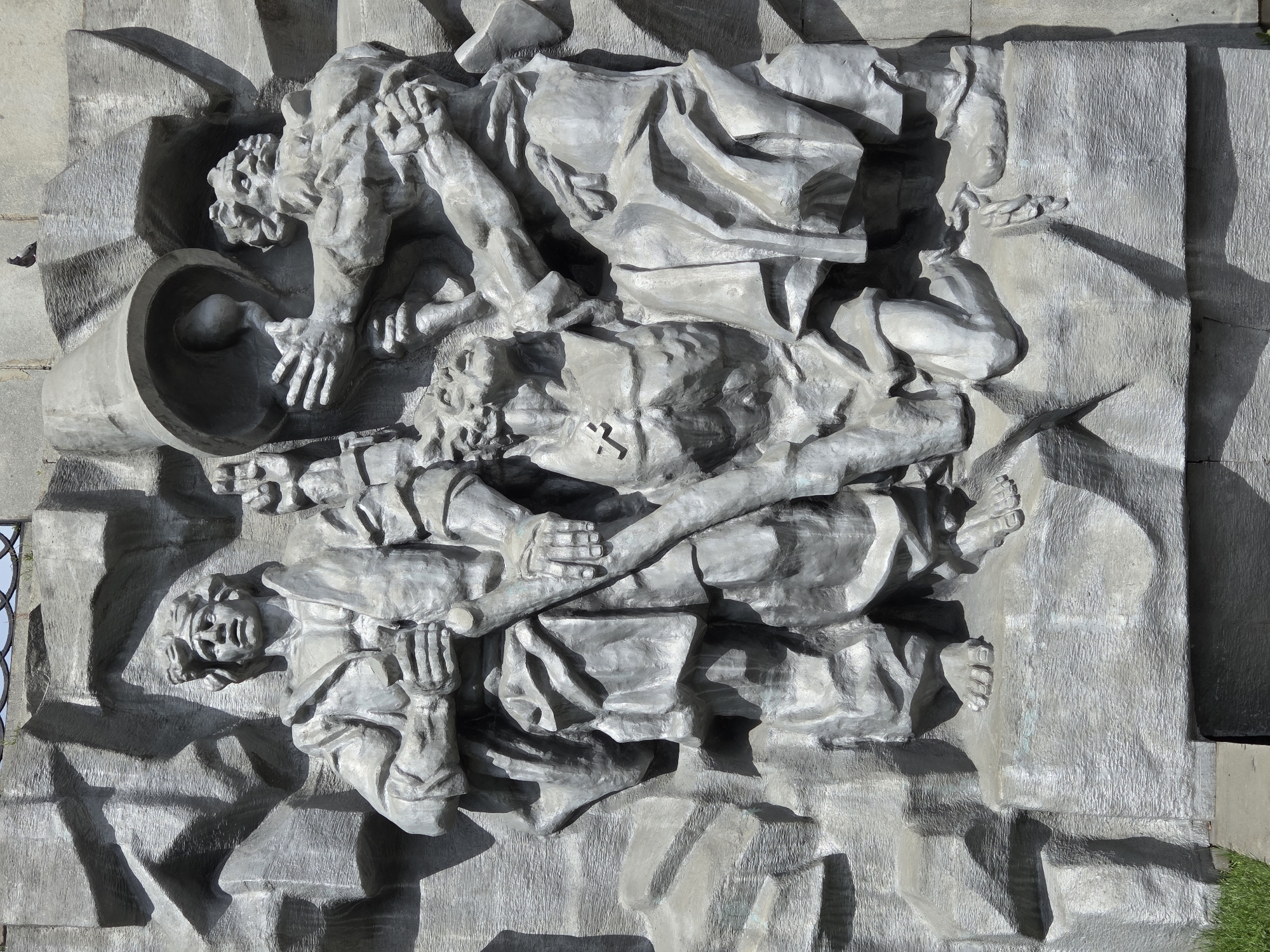 This is a medium shot of a stone bas-relief sculpture located in Yekaterinburg, Russia. The sculpture depicts three figures. On the left, a man stands with a determined expression, holding a staff. In the center, a kneeling figure with a cross on his chest looks upwards. To the right, another man stands, reaching towards a large inverted bell that is being held aloft by the kneeling figure and the man on the right. The figures are rendered in a stylized, somewhat rough manner, with sharp angles and dramatic shadows. The background of the sculpture is also made of rough, textured stone, suggesting a rocky or mountainous terrain. A small pigeon is perched on the upper right edge of the sculpture. The lower left corner of the image shows a patch of green grass.