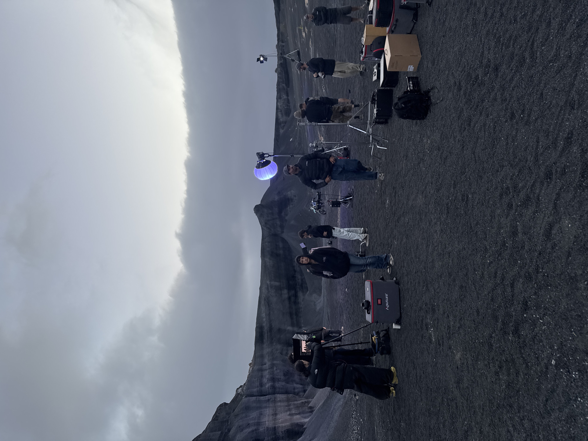 A film crew is working on location. The location is a black sand and rock landscape under an overcast sky. The crew is comprised of about 10 people, some standing around and some operating cameras and lights. There is a blue light on a stand. There are also camera cases and boxes of equipment visible. The precise location in Teguise, Spain, cannot be determined without more specific landmarks.