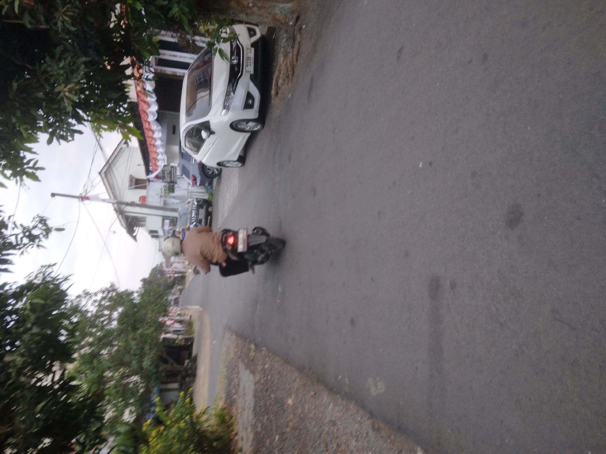 In Desa Pamijen, Kecamatan Sokaraja, Indonesia, a person wearing a light brown jacket and a white helmet is riding a black motorcycle away from the camera on an asphalt road. The motorcycle has a red taillight illuminated. To the right of the road, a white car is parked, and behind it, a grey car is also parked. A building with white siding and a red-trimmed roof is visible in the background, along with trees and overhead wires. The environment appears to be a street lined with houses and vegetation, with a slightly overcast sky.
