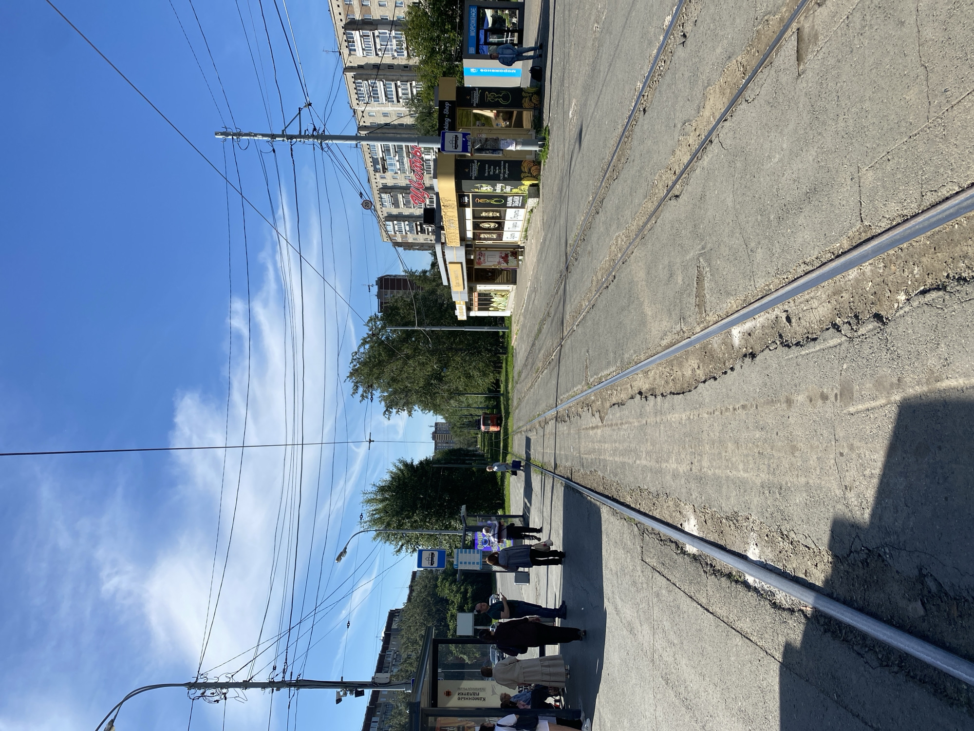 The image shows a tram line in Yekaterinburg, Russia, on a sunny day. In the foreground, tram tracks run down the center of a paved road. On the left, a tram stop shelter is visible with several people waiting. A small kiosk selling flowers and ice cream is located further down on the right side of the road. In the background, residential buildings and trees line the street, and a tram can be seen in the distance. The sky is blue with scattered white clouds.