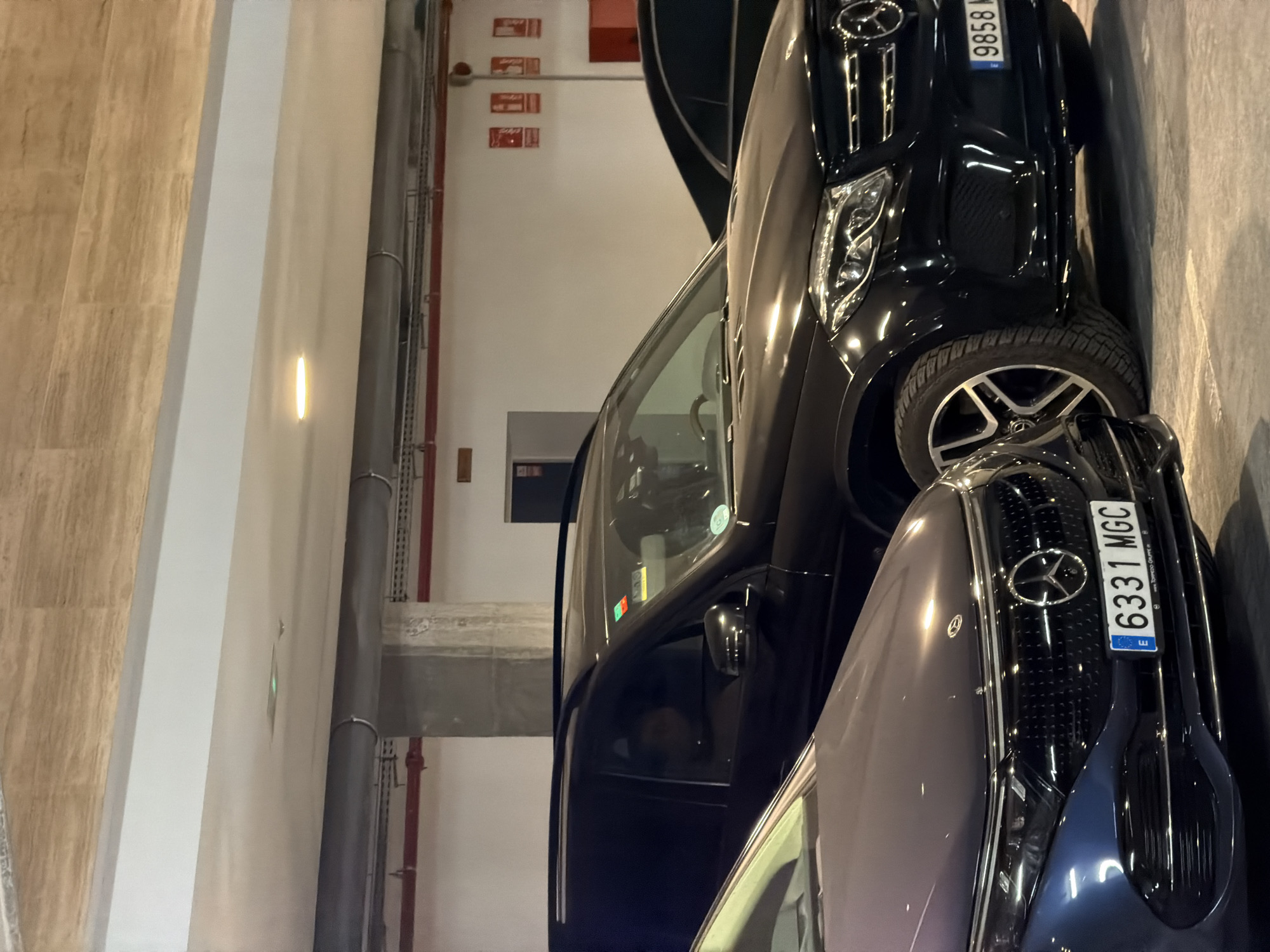 The image shows two Mercedes-Benz cars parked in an indoor parking garage. The car in the foreground, on the left, is dark blue and has the license plate "6331 MGC". The car behind it and to the right is black and has the license plate "9858 M...". The parking garage has concrete pillars and a white ceiling with pipes and lights. The scene appears to be in Finestrat, Spain, based on the context provided. There are no visible people interacting with the vehicles.