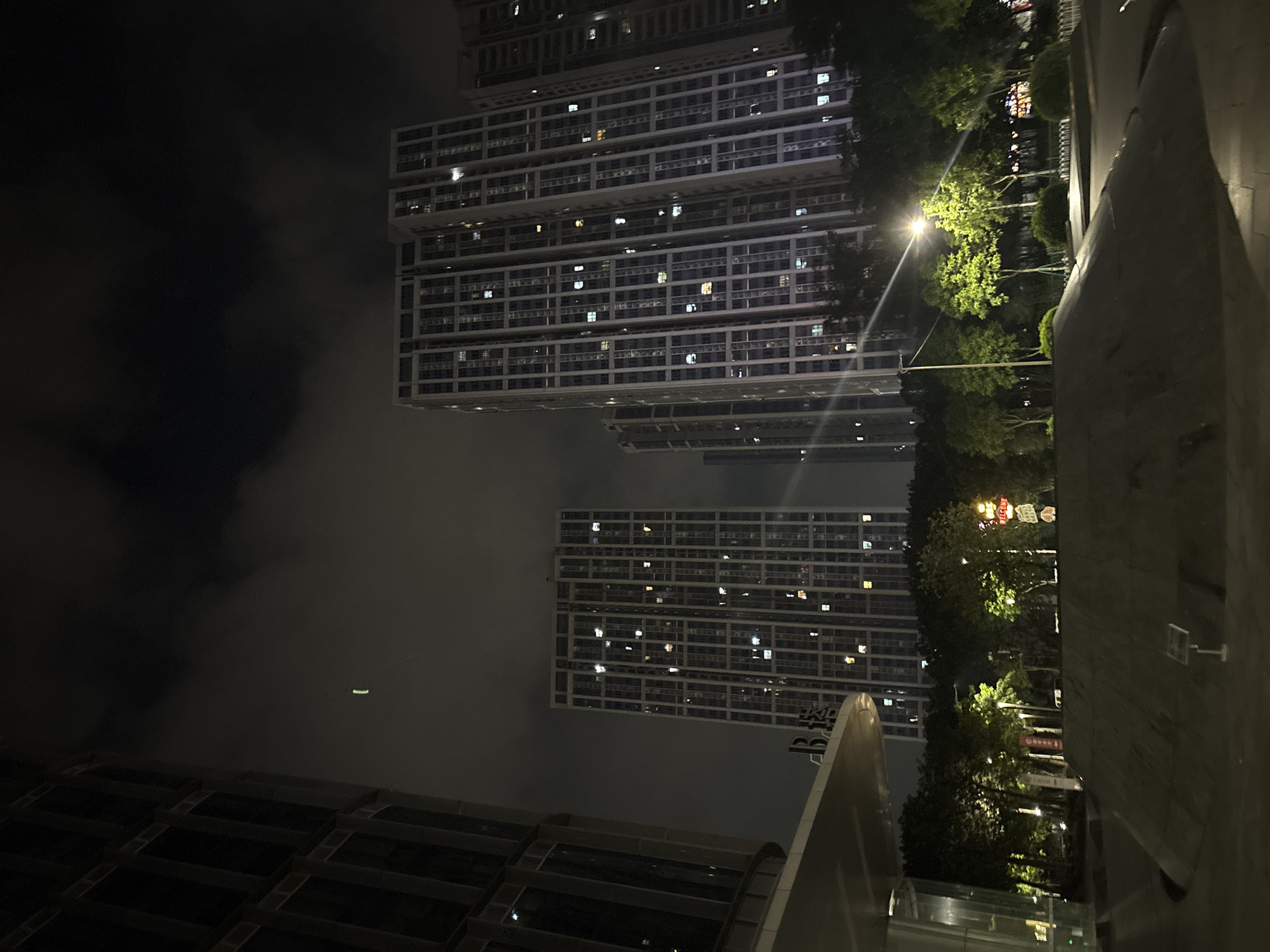 The image depicts a night scene in a modern urban environment, likely Haizhu District, China, characterized by towering residential buildings with illuminated windows. The perspective is from ground level, looking upwards towards the structures, which are partially obscured by dark, cloudy skies. A single streetlight is visible, casting a bright glow on lush green trees and other vegetation flanking a paved walkway or road.
The buildings are characterized by repetitive, grid-like facades, suggesting a high density of housing. The windows of these buildings show a scattering of lights, indicating that many residents are home. In the distance, a blurry sign with some colorful lettering is visible among the trees. A small, oblong green light is also visible in the sky, possibly a distant aircraft or a drone.
The overall atmosphere is somewhat muted due to the nighttime setting and cloudy weather, creating a sense of quiet urban life. The presence of modern architecture and the hint of signage suggest a developed urban area. The image captures a moment of ordinary evening life in a Chinese city, with the buildings standing as imposing, illuminated structures against the dark sky. There is no visible text that can be clearly deciphered.