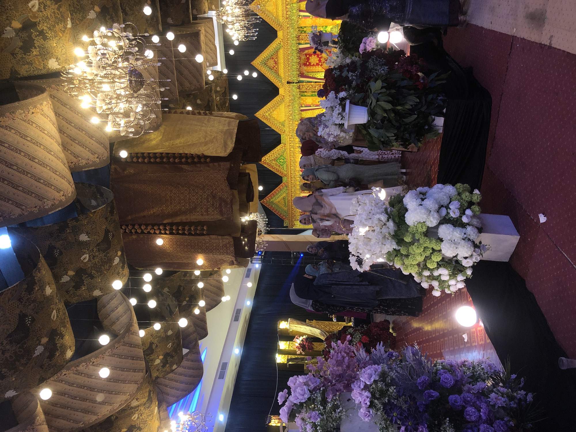 The image depicts an indoor event space, possibly a banquet hall, decorated for a celebration. The ceiling is adorned with draped fabric featuring a traditional Indonesian batik pattern, illuminated by numerous small, round lights and a large, ornate chandelier. In the background, a vibrant yellow and green structure with triangular peaks, likely a stage or decorative backdrop, is visible.
Several individuals, predominantly women wearing hijabs and modest attire, are present, interacting with each other and observing the surroundings. Flower arrangements, featuring purple, white, and red blooms, are strategically placed throughout the space, adding to the festive atmosphere. The floor is carpeted in a deep red hue. A single, bright spherical light emanates from the floor in the foreground.
Given the cultural context suggested by the attire and decorations, this scene is likely a wedding reception or a similar significant social gathering. If this event is located in Lhokseumawe, Indonesia, it would be taking place within a venue designed to accommodate such cultural festivities.