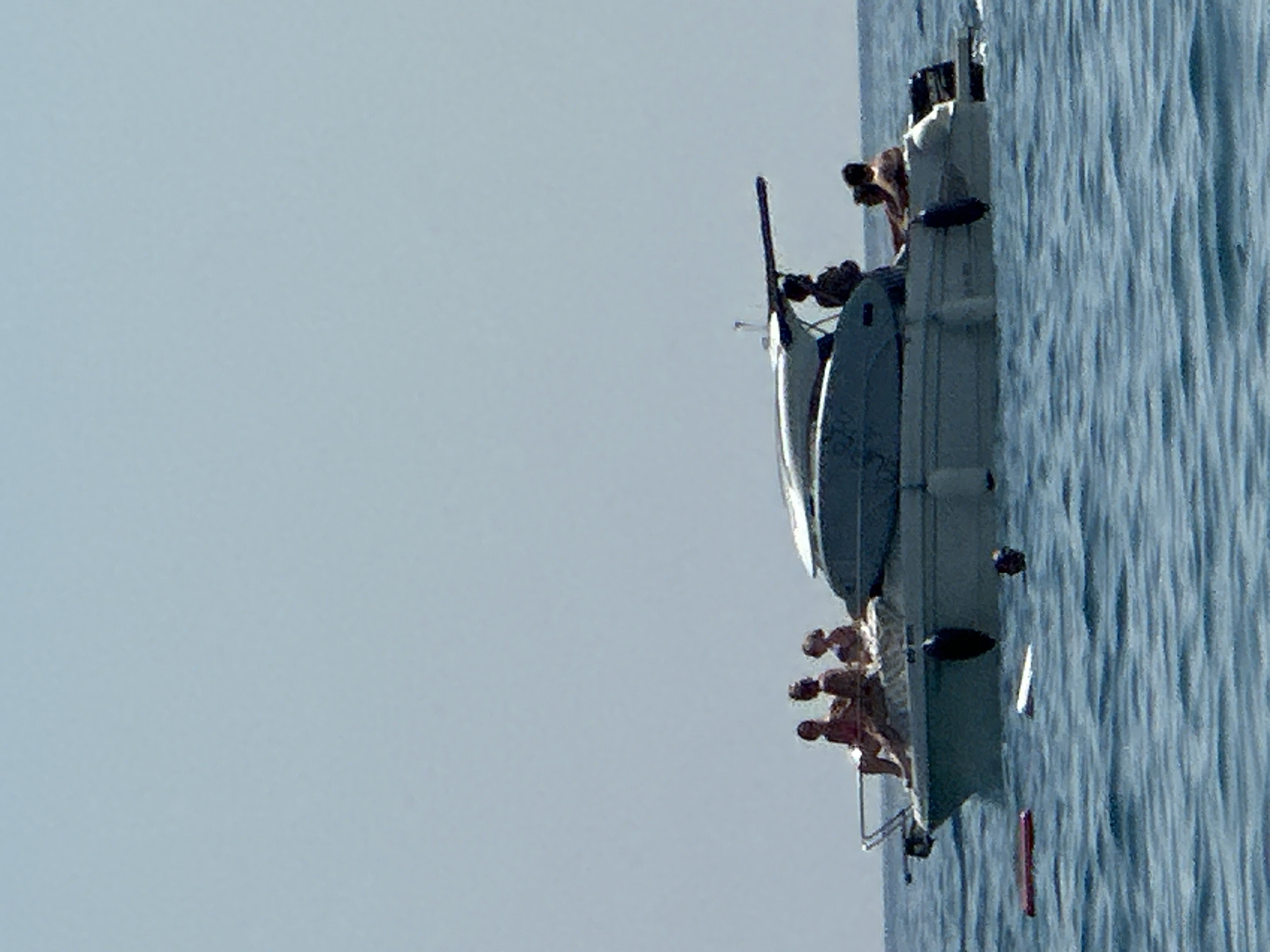 In the city of Boreti, Montenegro, on a clear, bright day, a white motor yacht is anchored in the blue, choppy waters of the Adriatic Sea. A group of six individuals are on board the vessel. Three people are seated on the front deck, appearing to be sunbathing. On the stern, another three individuals are also present, with two of them seated and one standing. The boat has a white cabin structure with a dark-colored canopy. In the water near the yacht, there are several floating objects, including what appears to be a red float and a white float, along with the head of a person in the water. The overall scene depicts a leisurely day on the water.