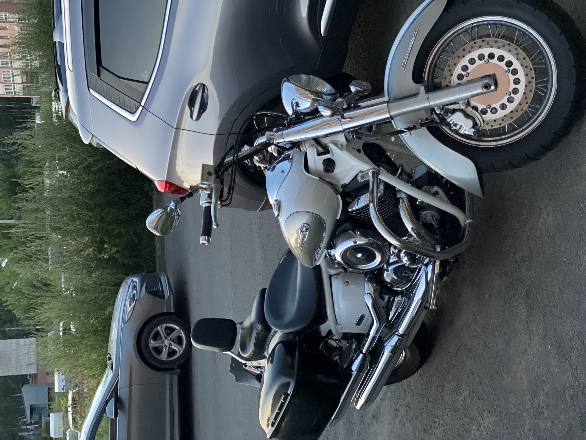 A silver motorcycle with black saddlebags is parked next to a silver SUV on an asphalt surface in a city environment. The motorcycle has a passenger backrest, chrome accents, and wire-spoke wheels. The SUV is partially visible, showing its rear section and a door handle. In the background, there is dense green foliage, a building with multiple floors, and a hint of a brick structure. The scene appears to be an outdoor parking area.
