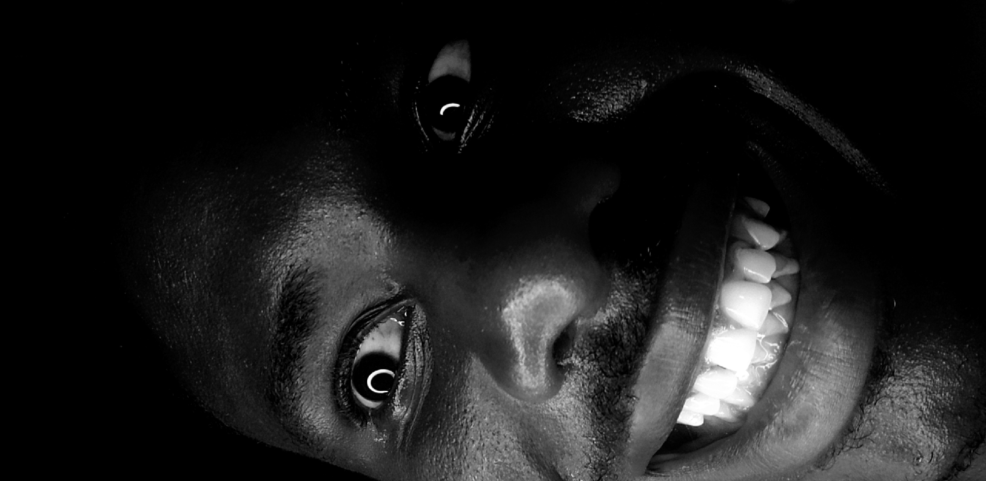A black and white close-up portrait of a person with dark skin. The person's head is tilted, so their eyes and teeth are prominently displayed. The person's mouth is open in a smile, revealing a full set of white teeth. The background is dark. The location of the image within Narayi, Nigeria cannot be ascertained.