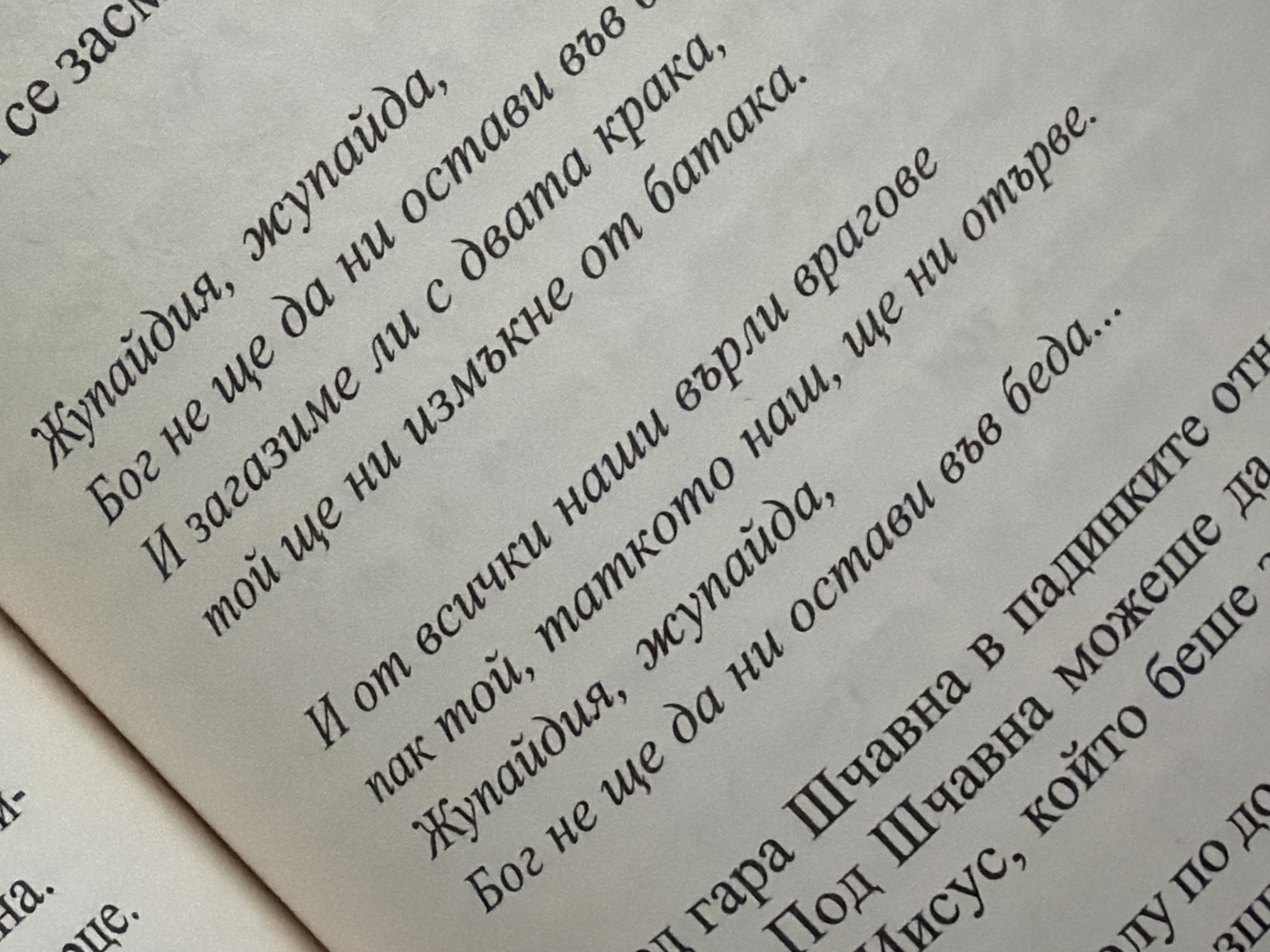 The image shows an open book with text in Bulgarian printed on its pages. The text is written in a stylized font and is angled on the page. The lighting suggests the book is illuminated by an overhead light source, casting subtle shadows. The environment appears to be indoors, possibly a home or library, with a soft, neutral background. There are no people visible in the image. The content of the text is religious or spiritual in nature, judging by phrases like "Бог не ще да ни остави" (God will not leave us) and "Исуc" (Jesus). The text is primarily in Cyrillic script.