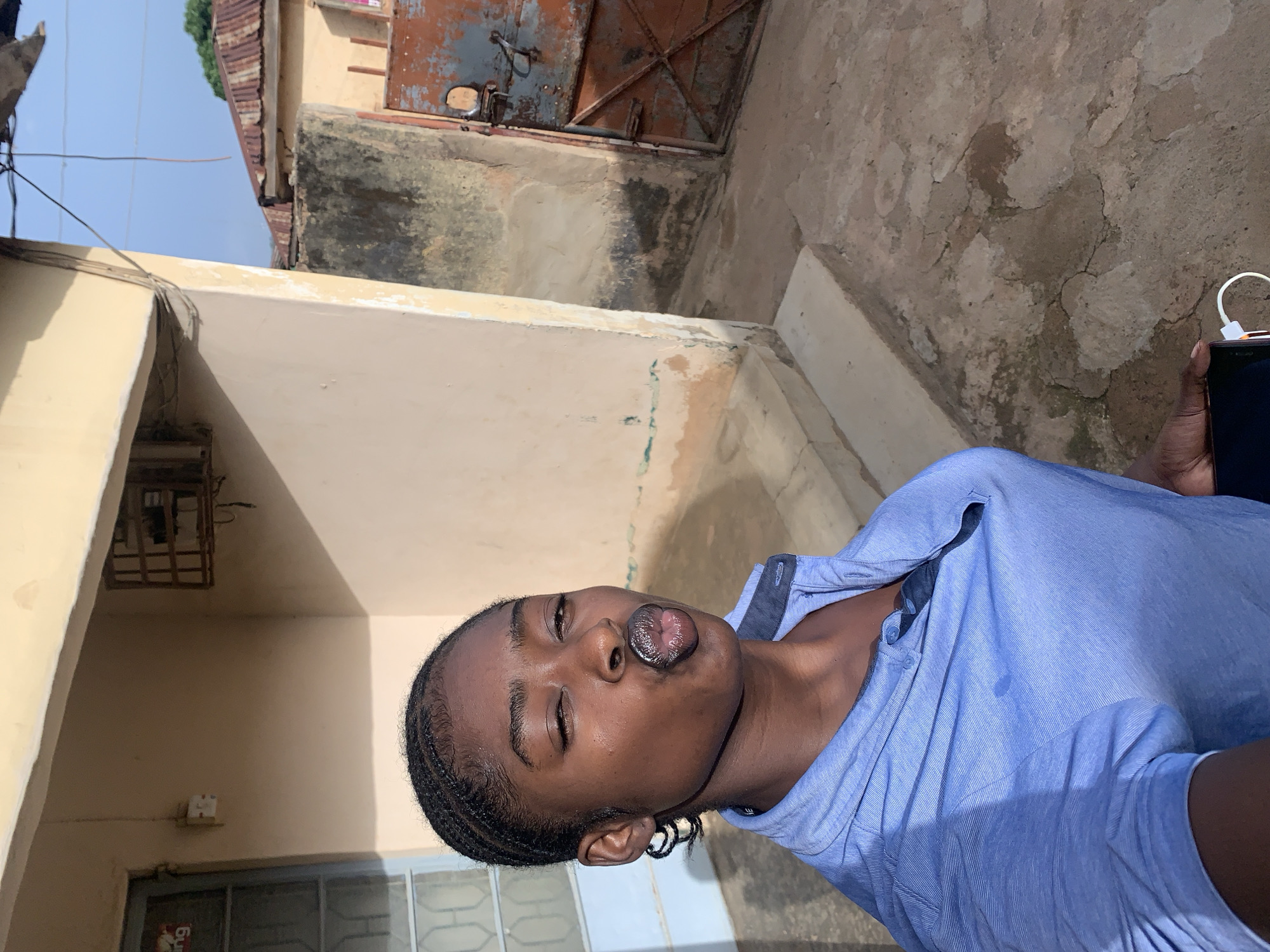 A young person with dark hair and skin is facing the camera with their lips pursed in a pouting gesture. They are wearing a light blue button-down shirt. The person is holding a smartphone with a white charging cable attached. They are standing in an outdoor location, with a cement wall and building visible behind them. The wall appears aged and stained, with variations in texture and color. A weathered metal door with a latch is also visible. The location appears to be Rafin Par, Nigeria, based on the environment. The lighting suggests it is daytime.