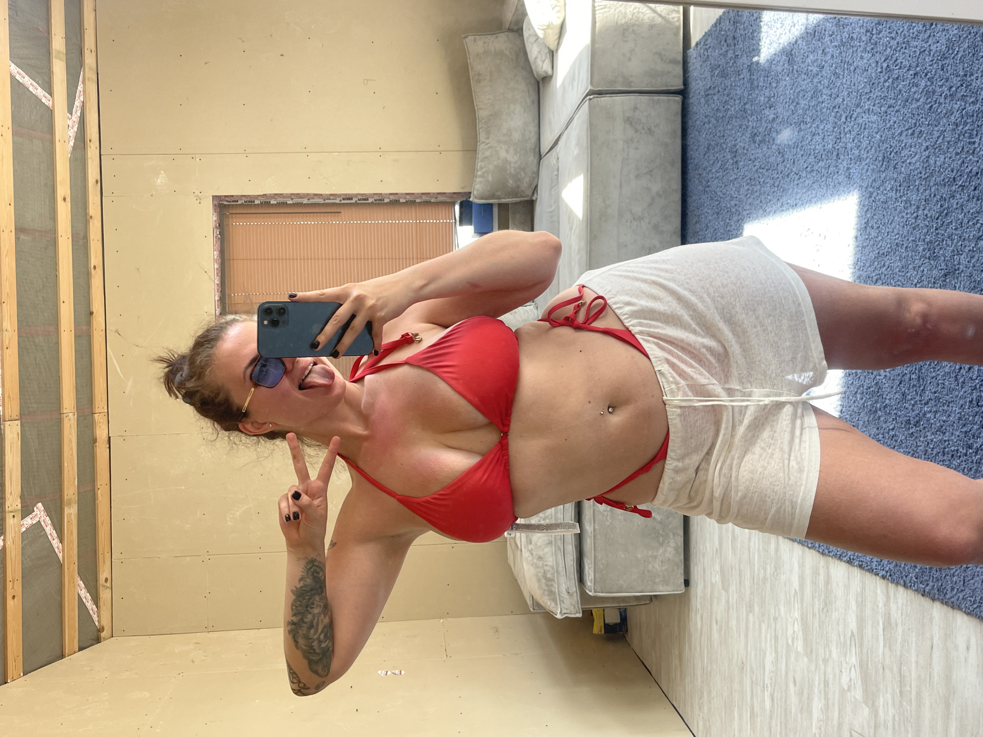 A woman is taking a selfie in a mirror. She is wearing a red bikini top and white shorts, and sunglasses. She is sticking her tongue out and making a peace sign with her left hand, which has a tattoo on the forearm. The background is an unfinished room. To the left is an unfinished wall. Behind the woman is a couch. A blue carpet is visible in the reflection. The location is Мосметаллоконструкция Онт, Russia.