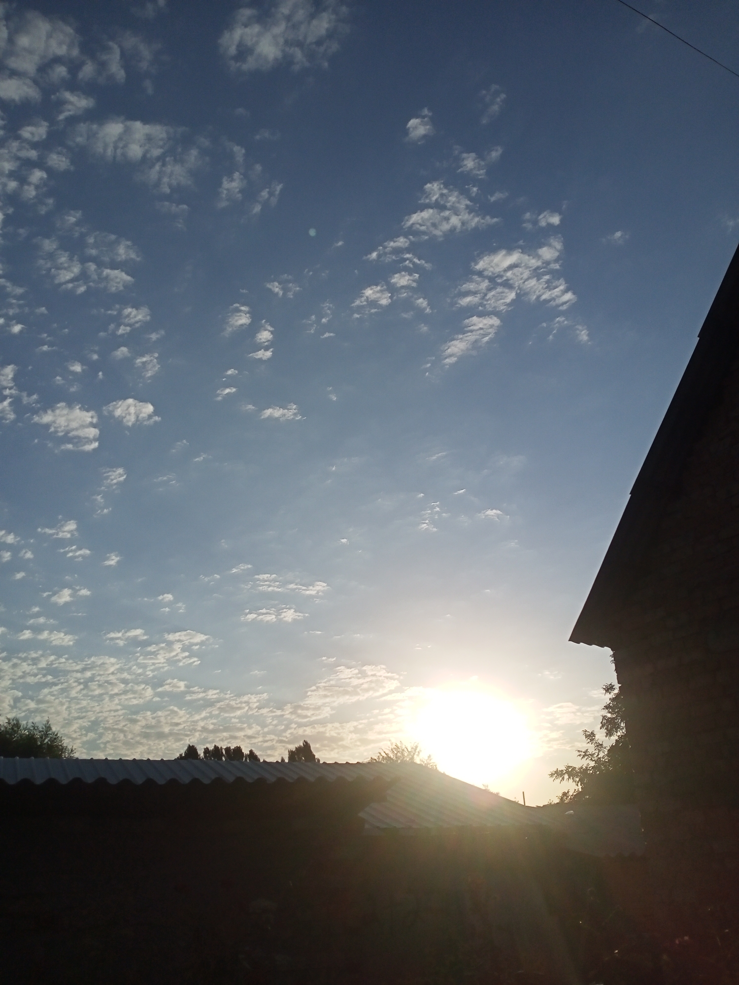 The image depicts a rural or suburban scene in Sokuluk, Kyrgyzstan, at either sunrise or sunset. The sky is a clear blue with scattered white, wispy clouds. The sun, a bright orb, is visible low in the sky, casting a warm glow and long rays that create a lens flare effect. In the foreground, a dark silhouette of a building with a corrugated metal roof is visible. To the right, another building, also in silhouette, shows a steeply sloped roofline and textured walls, suggesting it might be constructed of brick or stone. Trees and foliage are present as dark shapes against the brighter sky. The overall impression is one of a tranquil evening or morning in a non-urban setting.