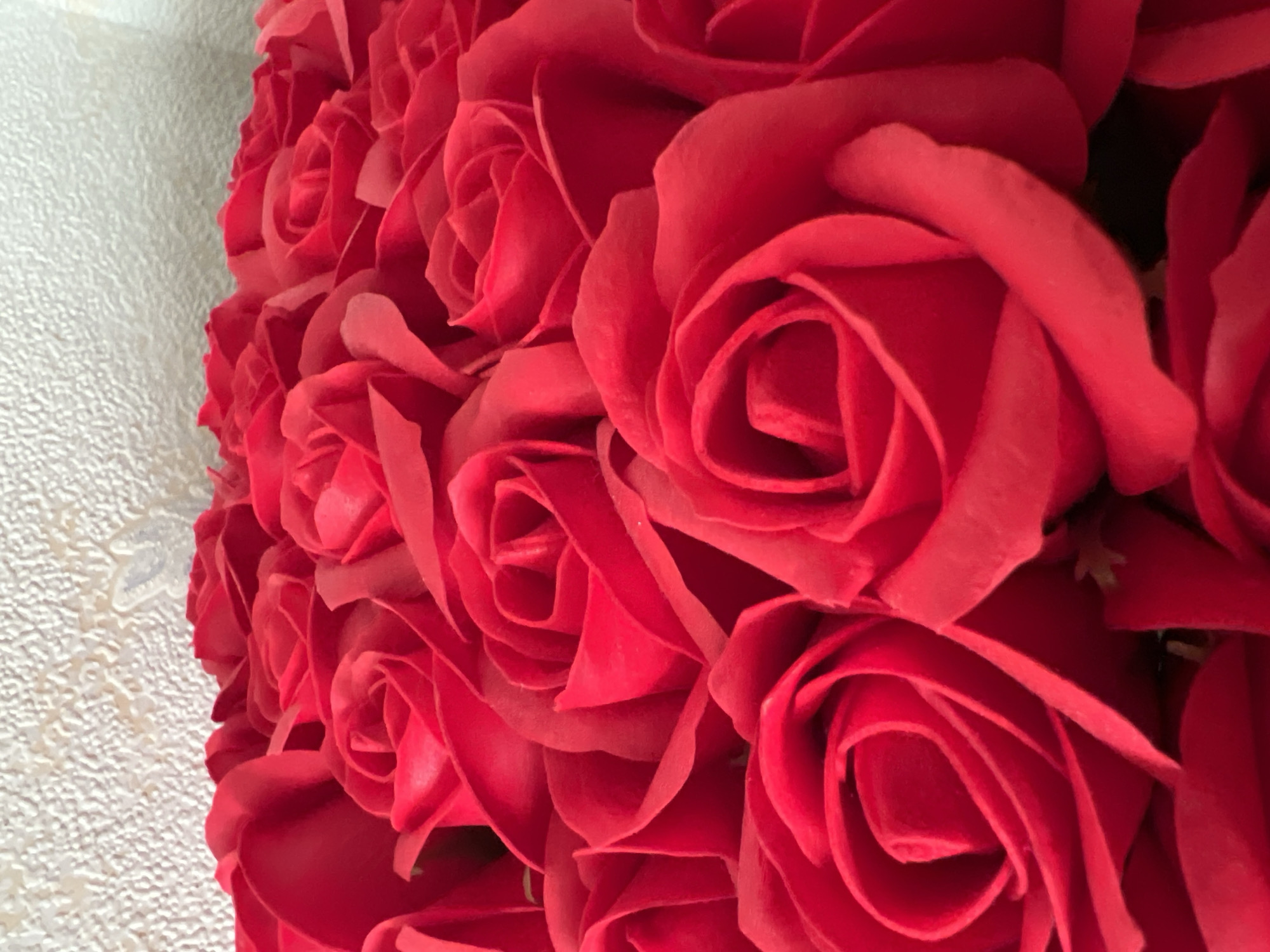 The image displays a close-up view of a bouquet of vibrant red roses. The roses are arranged densely, with many petals overlapping and obscuring the centers of some flowers. The background is a textured, pale surface, possibly a wall or fabric, which is out of focus. There is no indication of the location being Heorhiivka, Ukraine, nor are there any people, actions, or interactions depicted. The focus is solely on the texture and color of the rose petals.