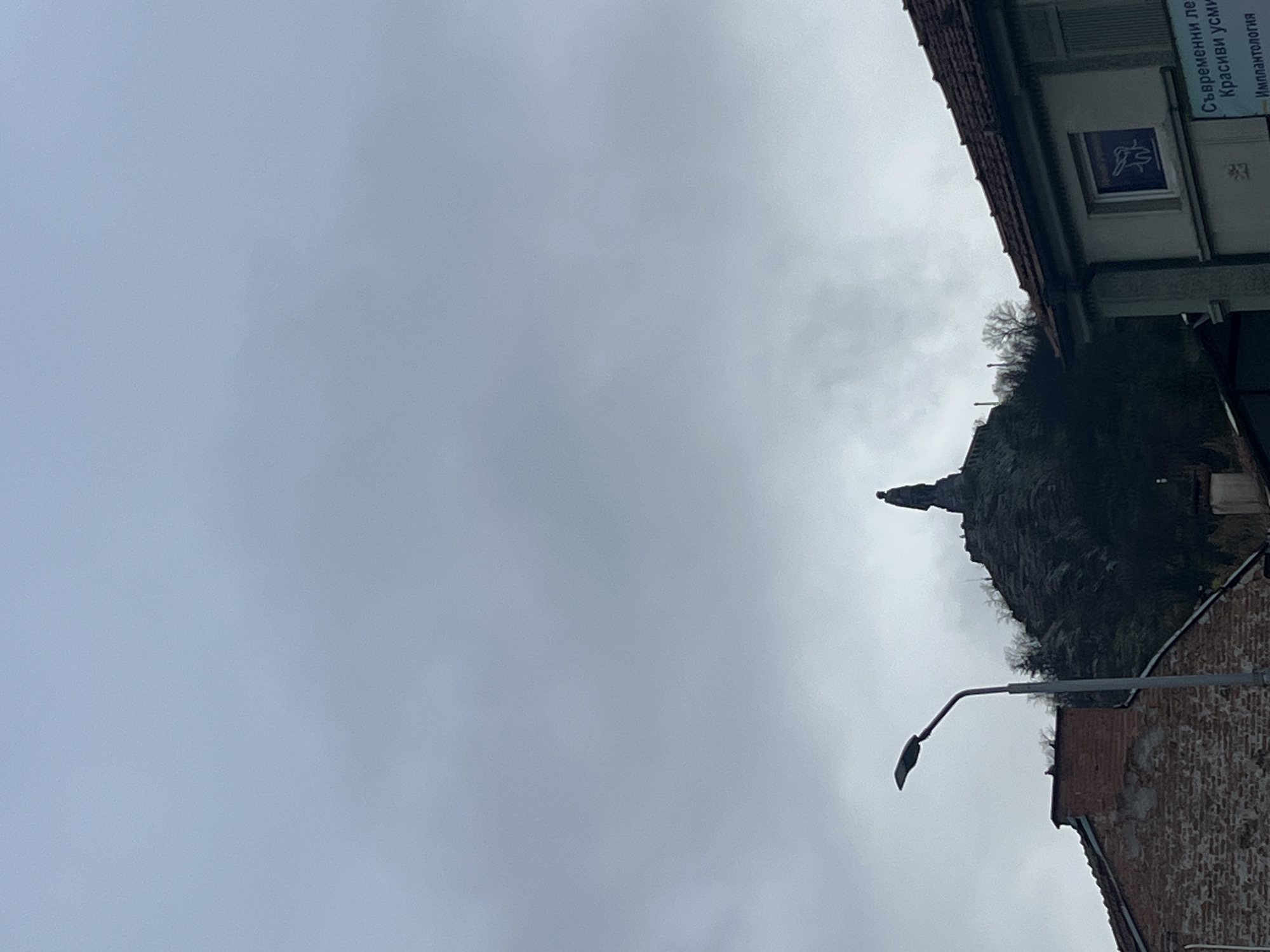 The image depicts a cloudy sky over Plovdiv, Bulgaria. A statue stands atop a small mountain covered in trees. A street lamp is visible in the lower right corner, along with a brick building. On the upper right, a building with signs written in Cyrillic is partially visible. The scene appears to be taken during the day.