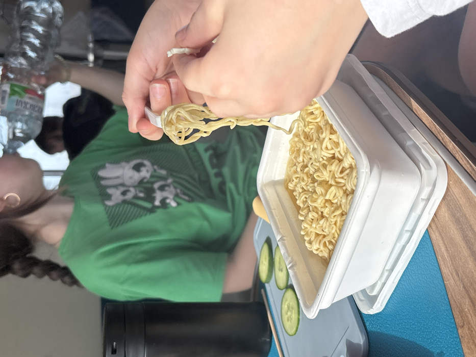 The scene depicts an indoor setting, likely a dining area, in Karmanovo, Russia. In the immediate foreground, a pair of hands is active; the left hand holds a white plastic fork, from which a portion of yellow instant noodles is lifted, while the right hand holds a small, irregular piece of white plastic, possibly packaging. A white rectangular container of instant noodles rests on a white plastic tray, situated on a blue mat. To the left of the noodles, three circular slices of green cucumber are arranged on a lighter blue surface. A tall, black cylindrical thermos or bottle stands upright to the far left. In the midground, partially obscured, a person with dark hair styled in braids, wearing a green t-shirt with a dark graphic print, is drinking from a clear plastic bottle with a white cap, which displays Cyrillic text on its label. A gold hoop earring is visible on this person's left ear. Another indistinct figure is partially visible in the blurred background. The setting includes a light brown wooden table surface.