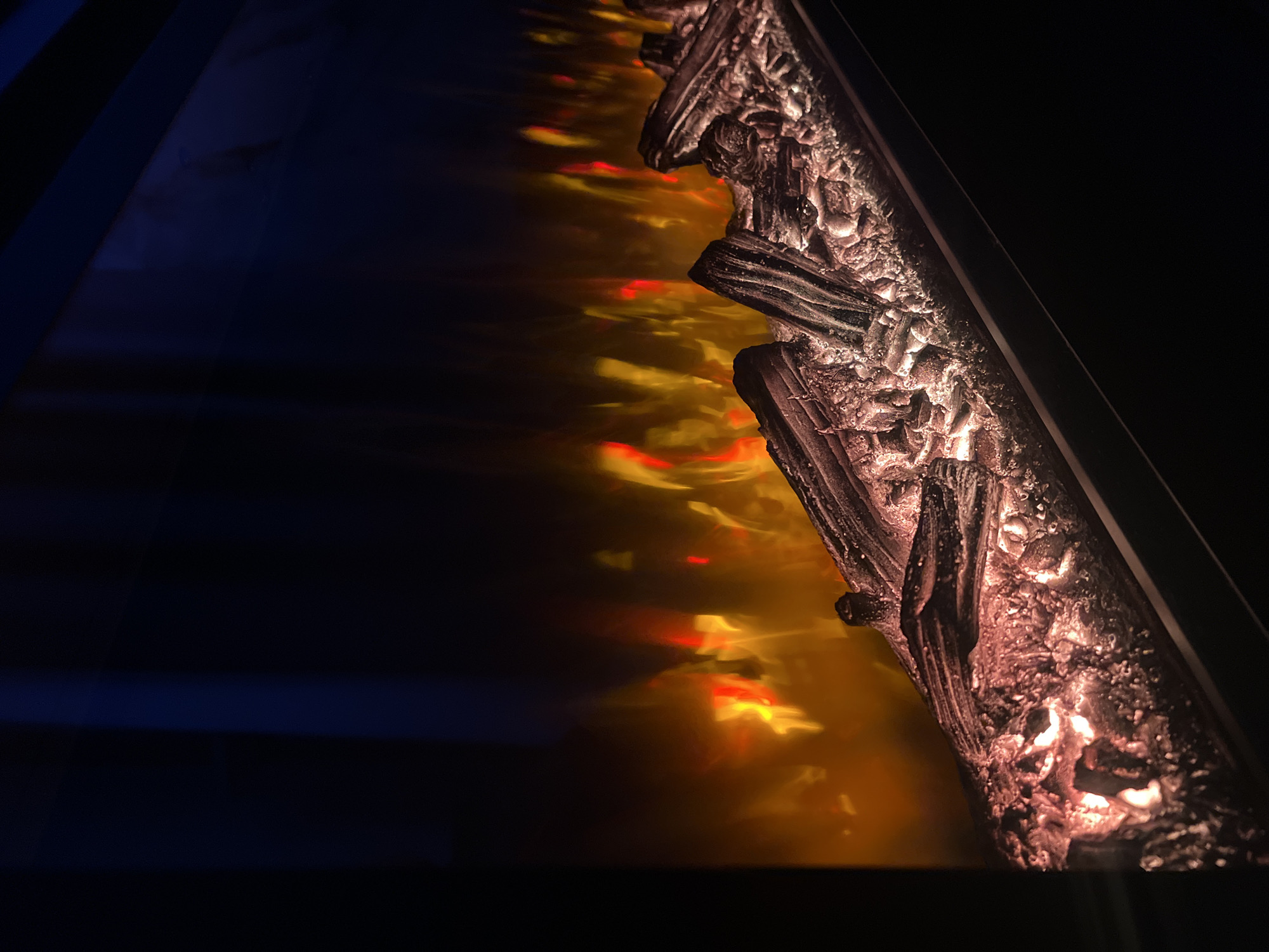 The image depicts a close-up of an electric fireplace, possibly indoors in Perm, Russia. The right side of the frame is dominated by the faux logs and a portion of the fireplace's structure, illuminated with warm, artificial light to simulate burning embers. The light casts an orange and red glow, creating the illusion of flames. The left side of the frame is dark, with shades of blue and black.