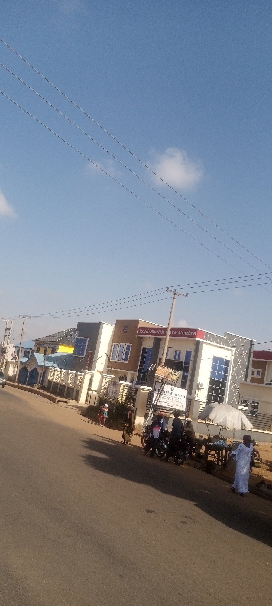 The image depicts a street scene in Sabon Tasha, Nigeria. The primary focus is on the "Roni-Healthcare Centre," a modern-looking building with brown and white accents, visible signage, and reflective windows. Adjacent to it is another building with a distinctive blue roof.
Several individuals are present on the sidewalk near the healthcare center. A woman in a white dress is walking toward the right of the frame, and a group of people is gathered around motorcycles parked in front of the building. A makeshift stall with a white umbrella is set up on the side of the road near the parked motorcycles. The road itself is paved and appears to be in reasonably good condition. Power lines are visible overhead against a clear blue sky with scattered clouds.
