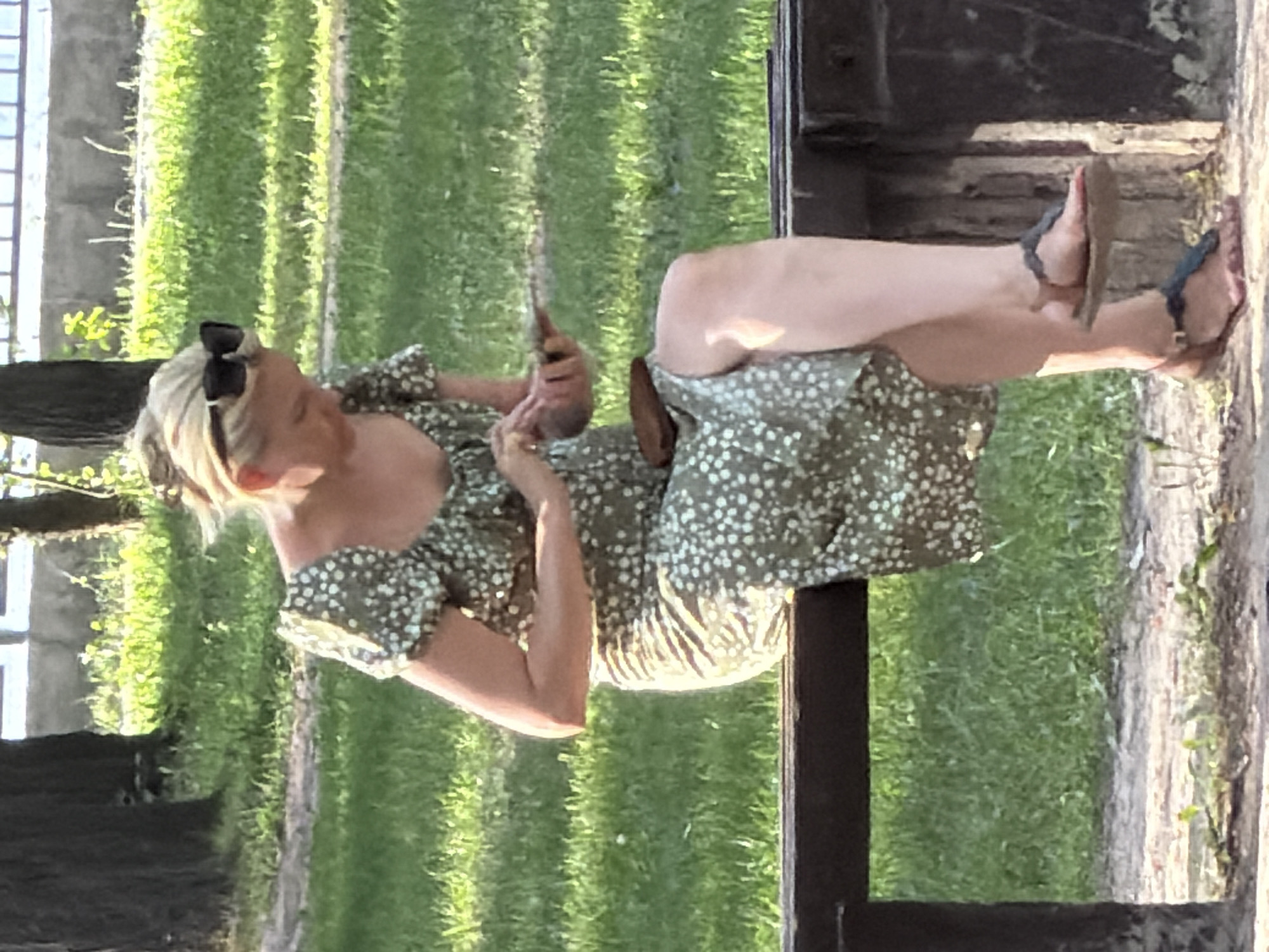 A blonde woman wearing sunglasses and a floral print dress sits on a dark wooden bench in a park. She is holding a mobile phone in her right hand, and her left hand is resting on her lap. The woman is wearing flat sandals. The background shows a grassy area with trees and a fence. The lighting suggests it is daytime. The location is Unknown, Unknown.