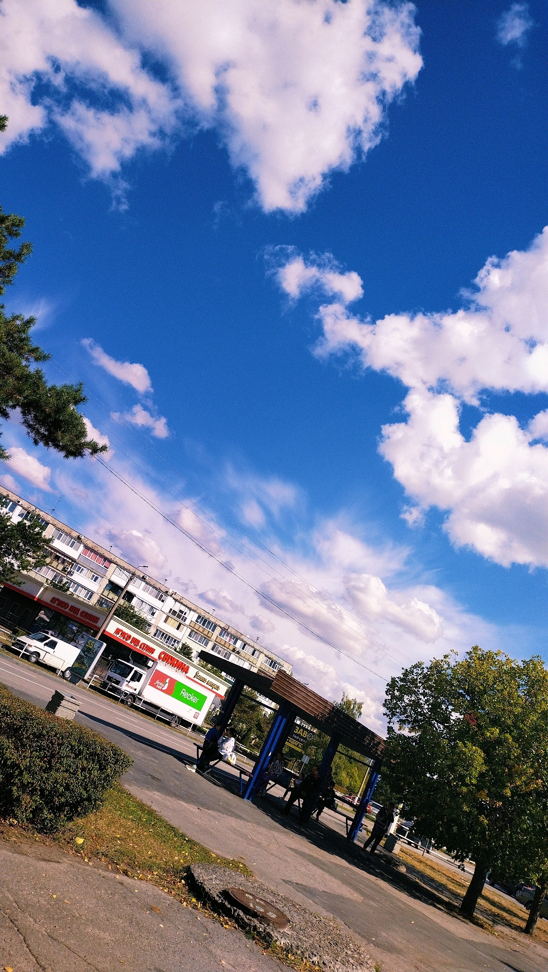 The image shows a street scene in Horishni Plavni, Ukraine, with a clear blue sky filled with white clouds. In the foreground, there is a sidewalk with a manhole cover. To the left, there is a green bush and a tree with fallen leaves on the ground. A bus stop structure with blue pillars and a brown roof is visible in the middle ground, with several people sitting and standing inside. Across the street, a long, multi-story apartment building is visible, with several shops on the ground floor. A white truck with a green "Flecker" logo is parked in front of one of the shops. A white van is also parked nearby. A large green tree stands to the right of the bus stop, with another tree visible behind it. The image is taken from a slightly tilted angle.