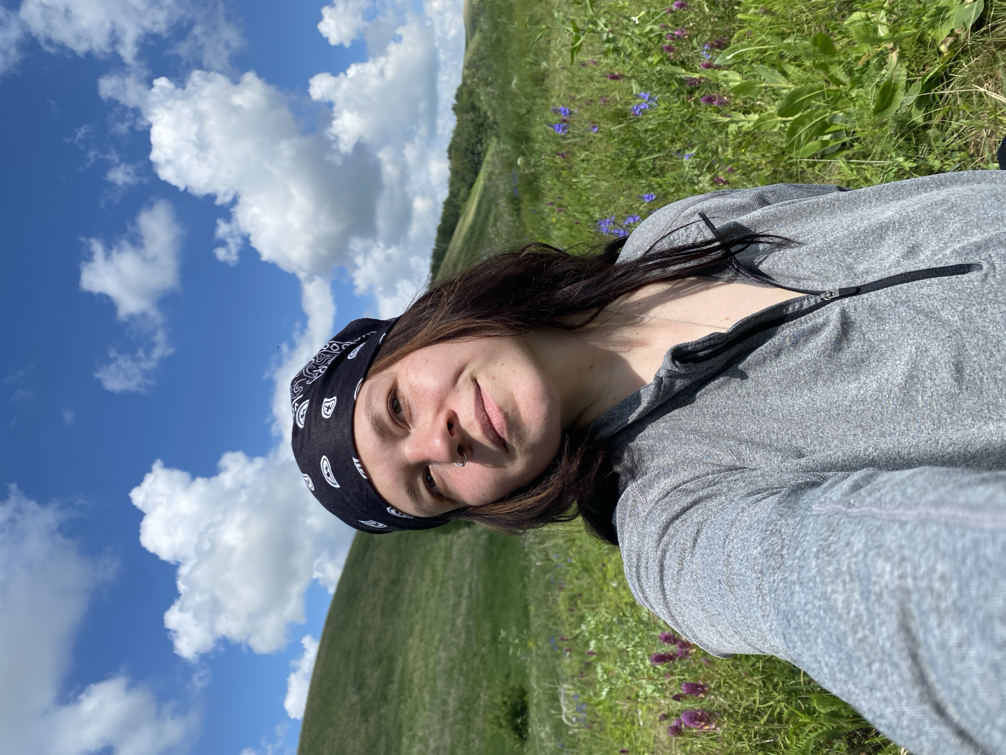 A young woman with dark hair, a black bandana, and a nose ring smiles at the camera while taking a selfie outdoors. She wears a grey zip-up jacket and a necklace. The background features a lush green hilly landscape with scattered purple and blue wildflowers under a bright blue sky with fluffy white clouds. The location is identified as Bogoslovka, Russia.