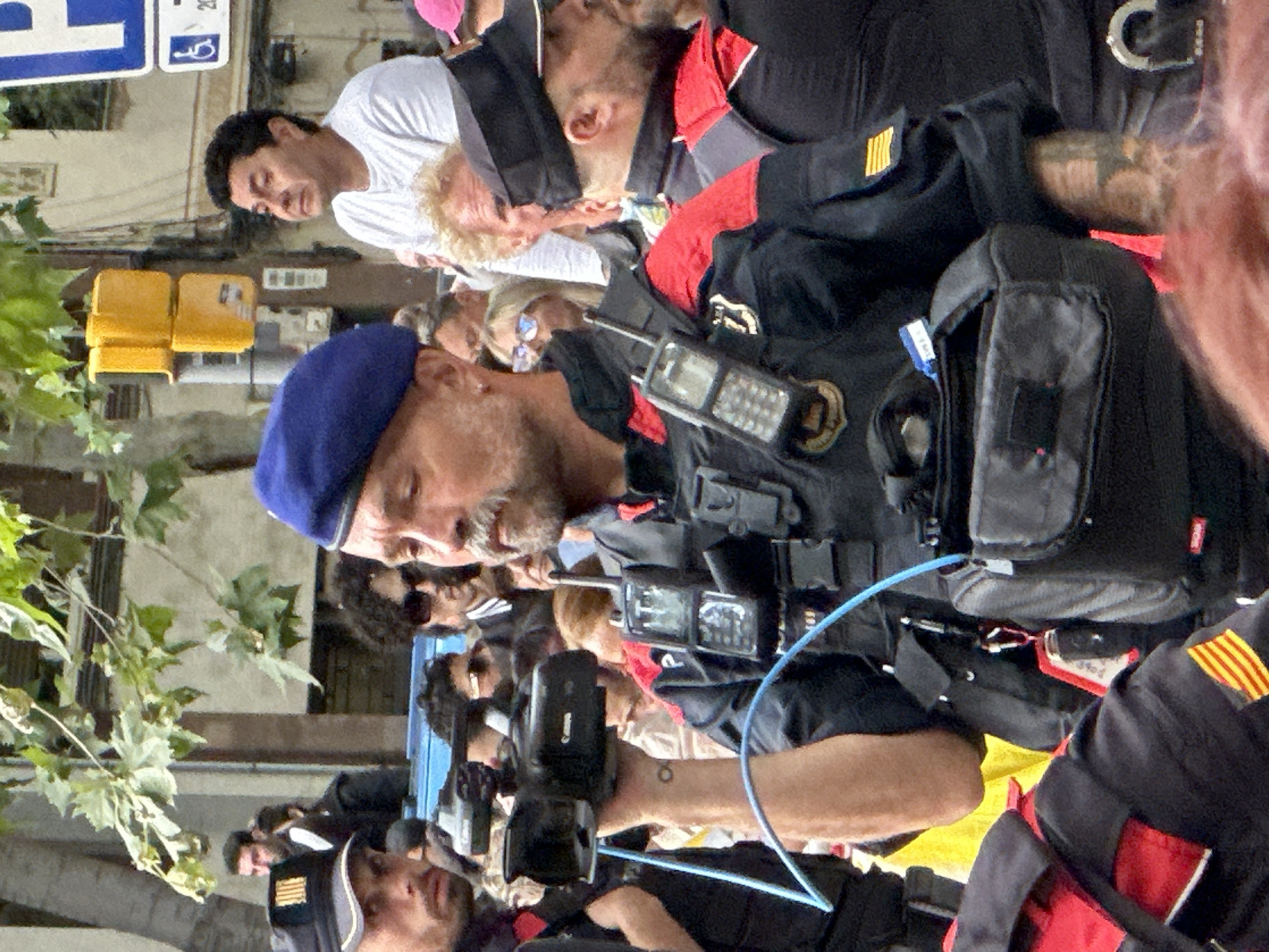 A man in a blue beret and tactical vest, holding a camcorder, stands in the foreground of an outdoor scene. He has a mustache and appears to be filming. Behind him, other individuals are visible, some wearing similar tactical gear, suggesting a public event or security detail. A traffic light with yellow signals is mounted on a pole to the left, and a building facade is visible in the background. The presence of Catalan flags on some of the vests indicates a location in Catalonia, Spain. The overall atmosphere suggests a public gathering or demonstration in Barcelona, Spain.
