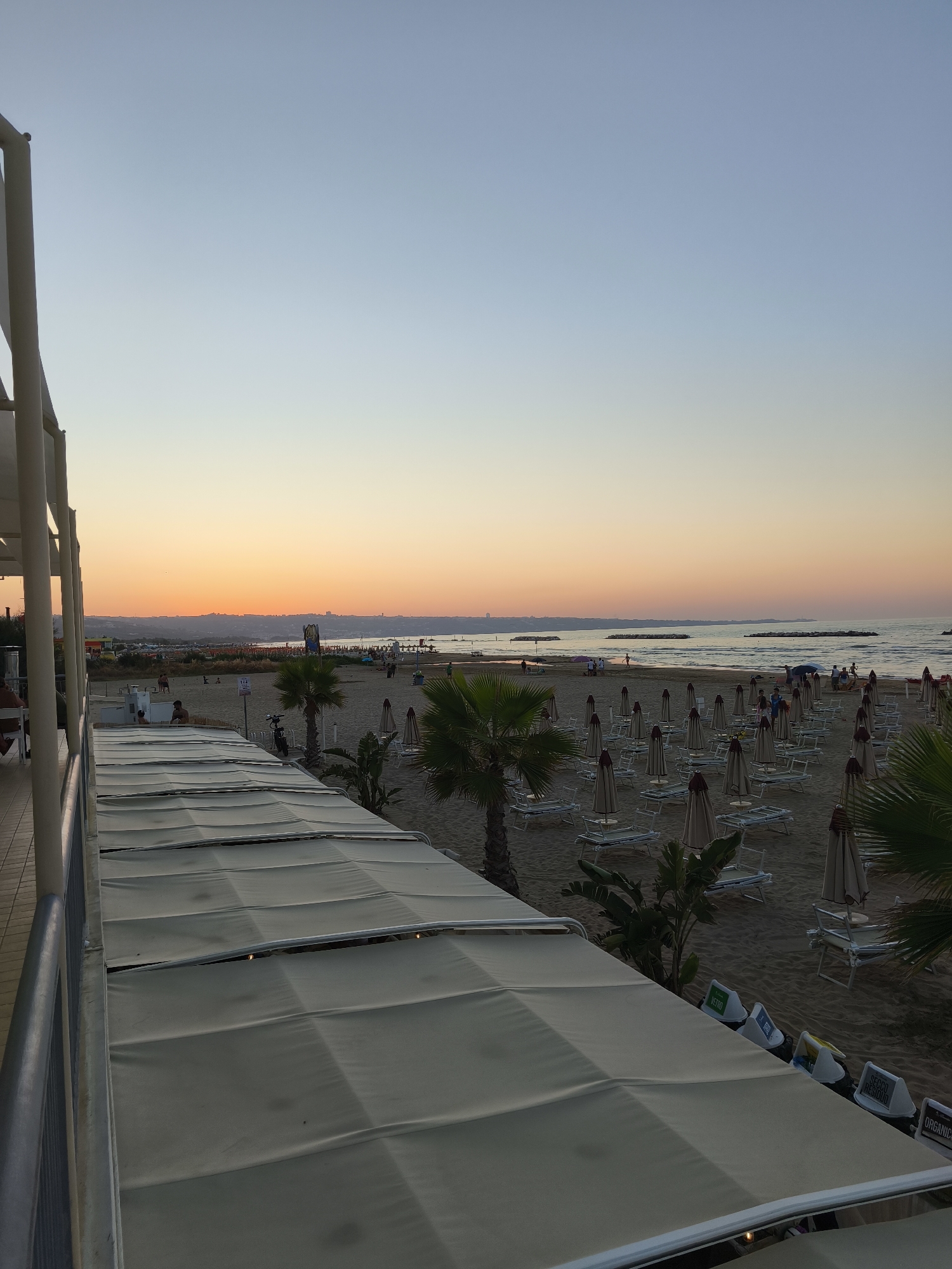 This is a wide-angle, eye-level photograph of a beach in San Salvo, Italy, at sunset. The sky transitions from a pale blue at the top to a warm orange and yellow glow near the horizon, indicating the setting sun. A distant shoreline with buildings is visible. The beach itself is sandy and dotted with numerous closed beach umbrellas and lounge chairs, many arranged in neat rows. Several palm trees and other tropical foliage are interspersed among the seating.
In the foreground, a portion of an outdoor seating area with a retractable awning is visible, supported by white pillars. A metal railing runs along the edge of this seating area.
On the beach, a few people are scattered about, some walking, others standing. There are signs and what appear to be recycling bins near the awning. The sea is calm, with gentle waves lapping the shore. The overall atmosphere is peaceful and serene, capturing the end of a day at the beach.