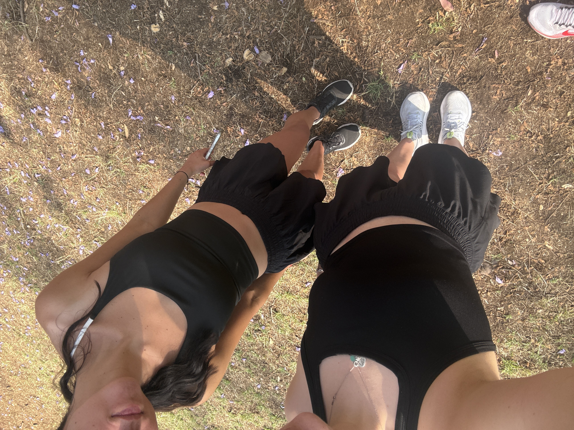 Two women in black athletic attire are standing on an area of sparse grass and bare earth scattered with purple blossoms, likely Jacaranda flowers, a common sight in Mexico City. The woman on the left wears a black sports bra and matching shorts, black running shoes and is holding a cell phone in her left hand. The woman on the right wears a similar outfit with white athletic shoes. The camera is held at eye level and angled down capturing the subjects from a selfie perspective. The presence of Jacaranda blossoms suggests the image was taken in Mexico City, Mexico.
