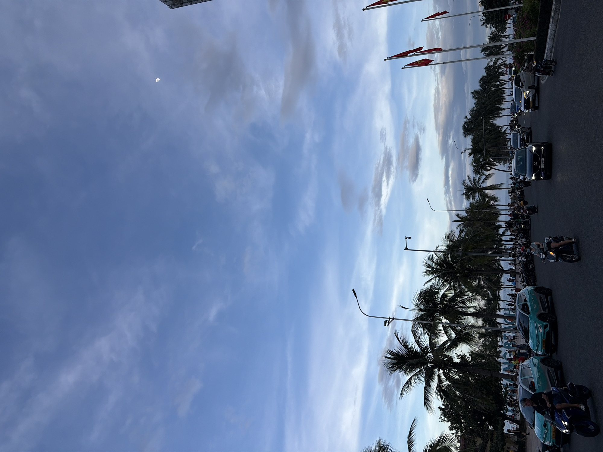 The image captures a street scene in Da Nang, Vietnam, likely during twilight or early evening. The sky is a prominent feature, displaying a gradient of blues with streaks of white clouds and a visible crescent moon in the upper right quadrant. A line of palm trees stretches across the middle ground, obscuring a beach and the ocean beyond. On the street, a moderate flow of traffic is present, including cars, motorcycles, and scooters. Several vehicles are light blue, suggesting a common rental or ride-sharing service. People are visible riding motorcycles and scooters, some wearing masks. In the background, numerous flags, predominantly red with a yellow star, are flying from poles along the roadside, indicating a public or official area. The overall atmosphere suggests a coastal city setting with ongoing urban activity.
