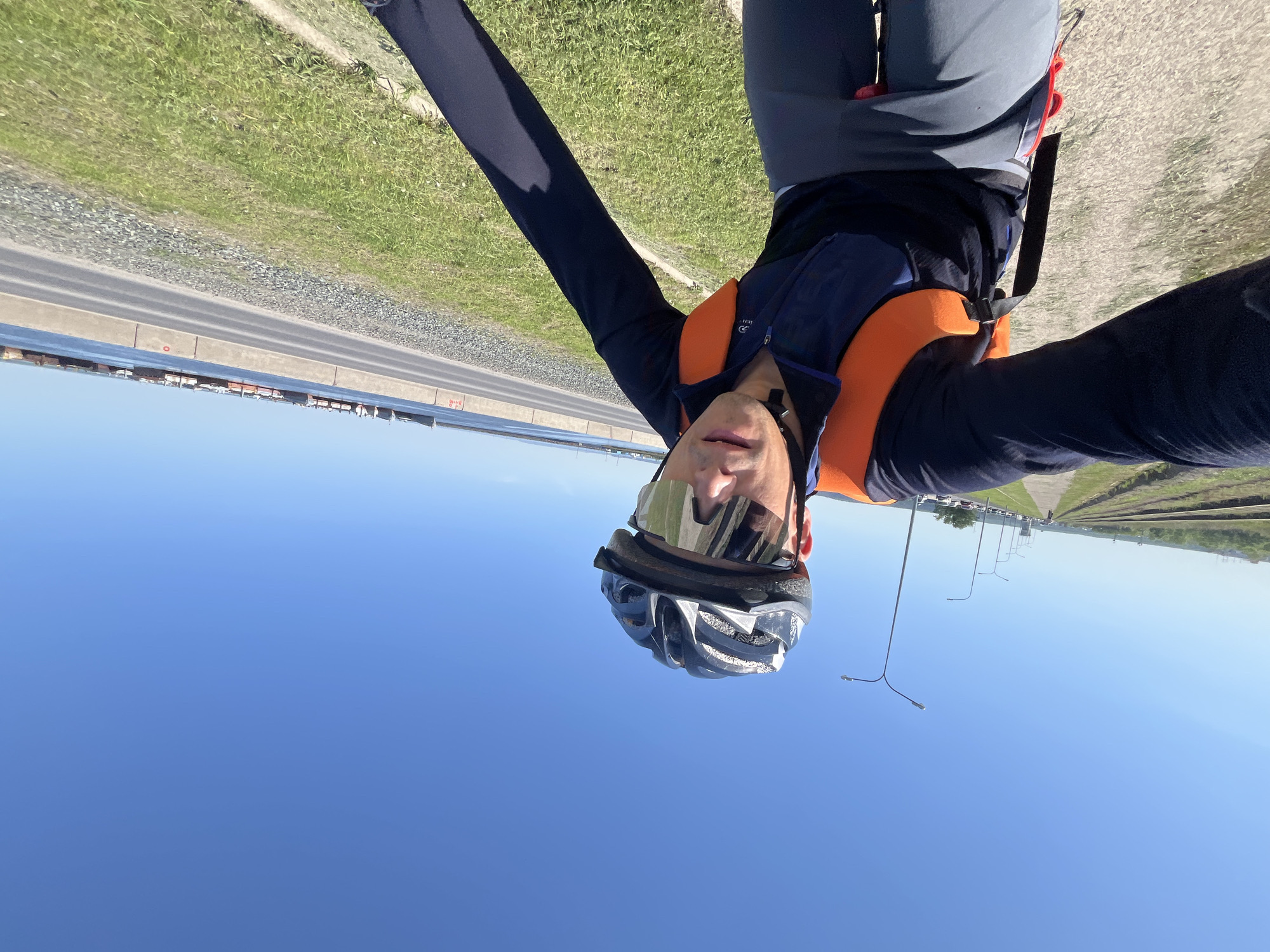 A selfie taken by a person wearing a helmet, sunglasses, and what appears to be a life vest. The background reveals a coastal view with a road and some industrial structures in the distance. The scene suggests an outdoor setting, possibly near a body of water and a transportation route, potentially taken in the city of Chaikovsky, Russia.