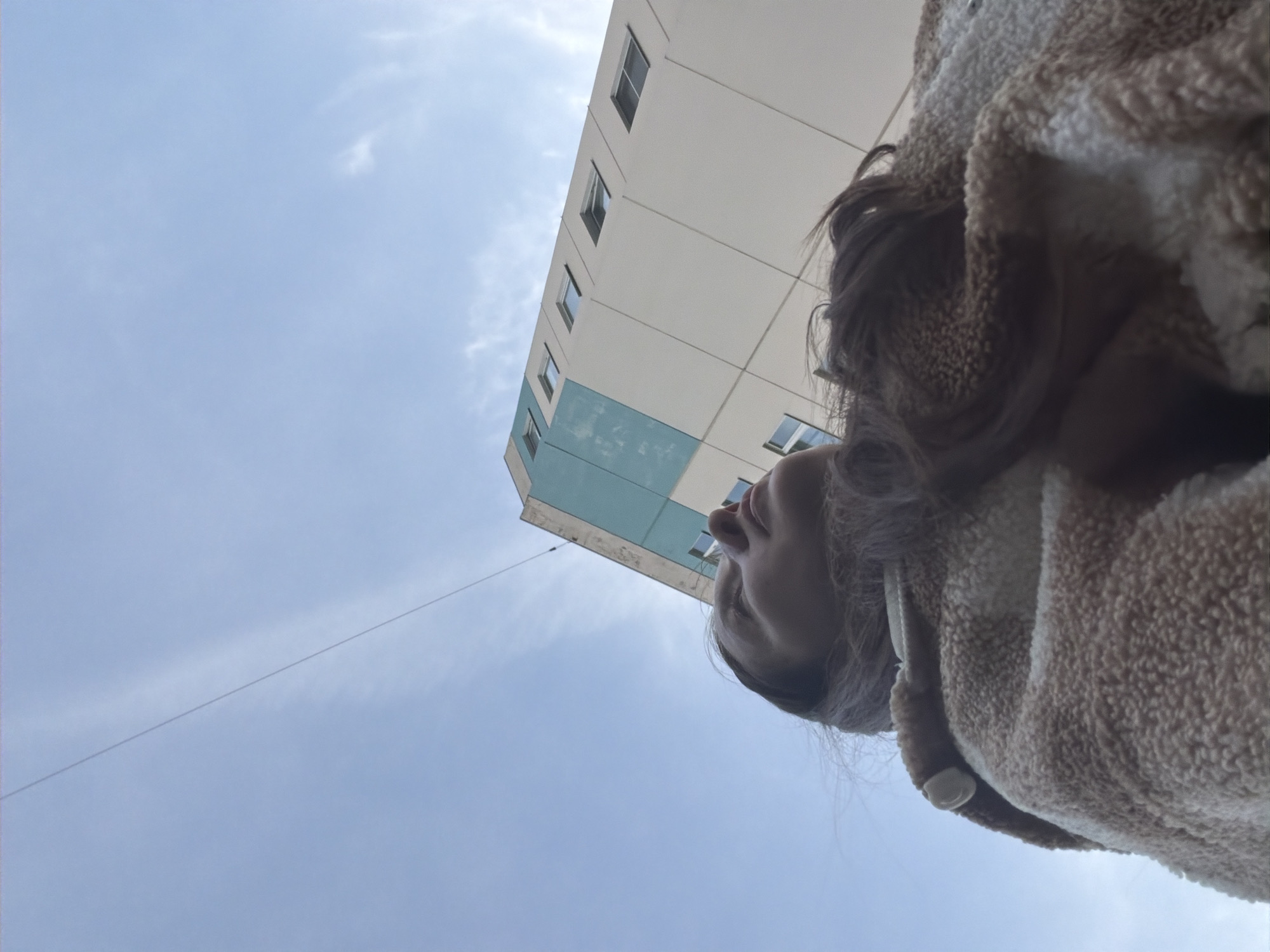 A low-angle shot depicts a person looking up at a white building with rows of windows against a cloudy sky. The building has a teal section on its corner. A thin wire stretches across the sky to the left of the building. The person is wearing a fuzzy brown jacket with the hood up, obscuring most of their face. The location is identified as Kolbino, Russia.