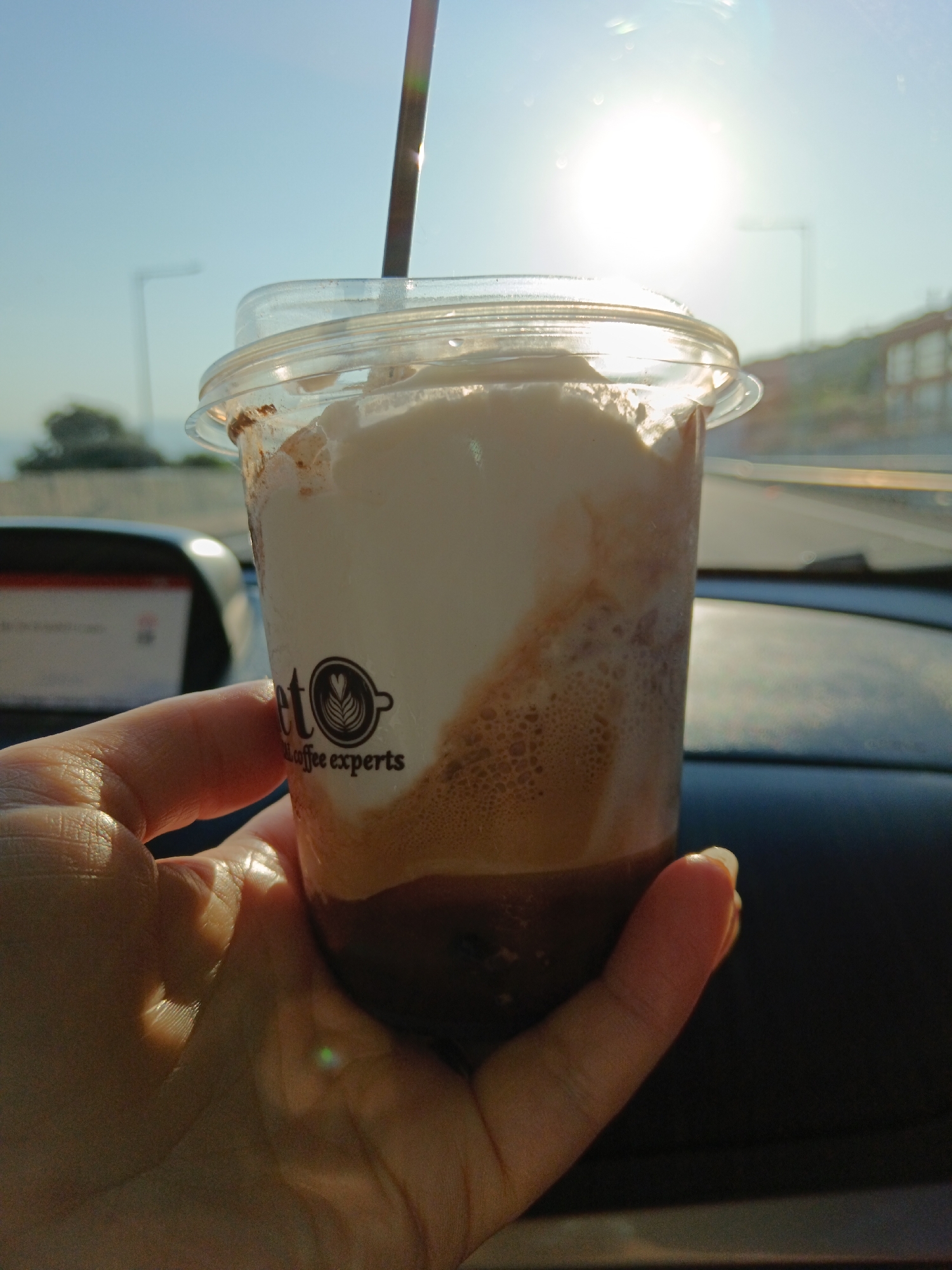 A close-up shot shows a hand holding a clear plastic cup filled with a layered beverage, possibly a coffee drink with foam or cream on top, and a dark liquid at the bottom. The cup has a black logo with text that appears to read "coffee experts". The background suggests an outdoor environment with a bright sun, possibly during daytime, with hints of a road and buildings in the distance. The location is identified as the Municipal Unit of Rio, Greece, indicating a scenic backdrop or a travel context.