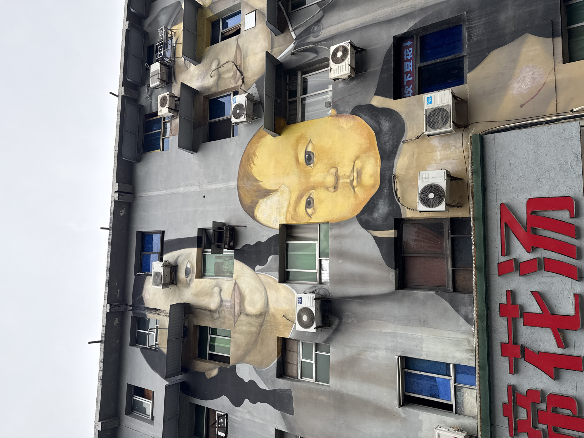 A tall building in Jiulongpo District, China is covered with a mural depicting the faces of two people, a child and a woman. The building has multiple windows with mostly open or closed gray shutters, and several air conditioning units are mounted on the exterior walls. A large red sign with Chinese characters is partially visible on the right side of the building. The sky is overcast.